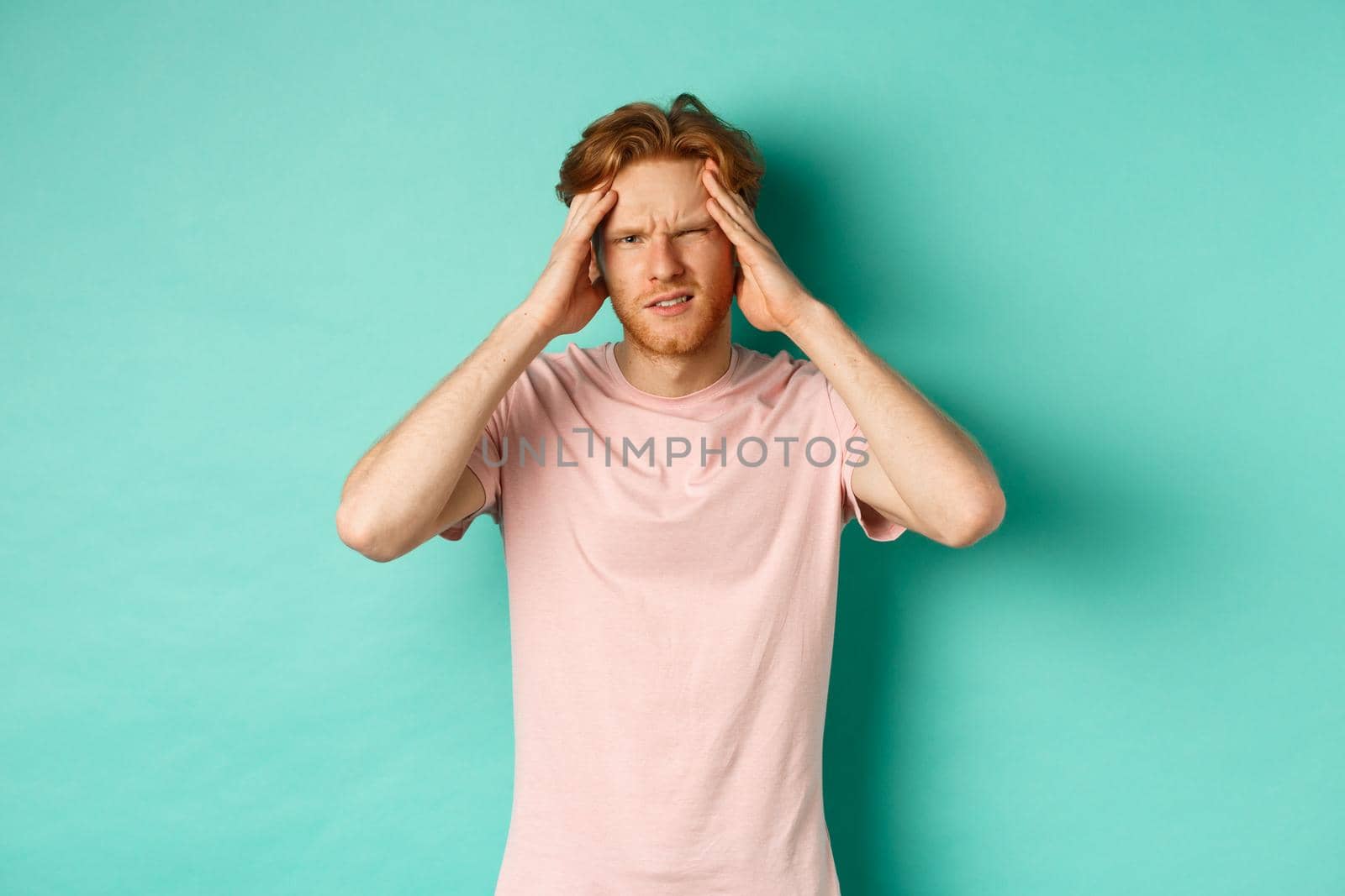 Young redhead man touching head and looking dizzy, feeling headache or migraine, standing in t-shirt over mint background.