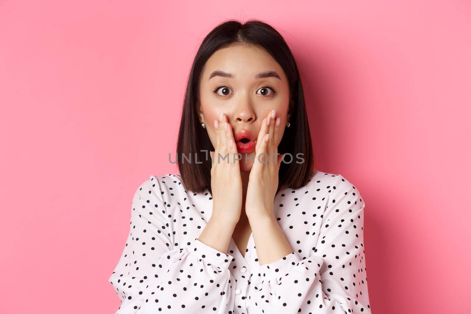 Close-up of surprised and amazed asian woman staring at camera, gasping impressed, holding hands near mouth, standing over pink background.