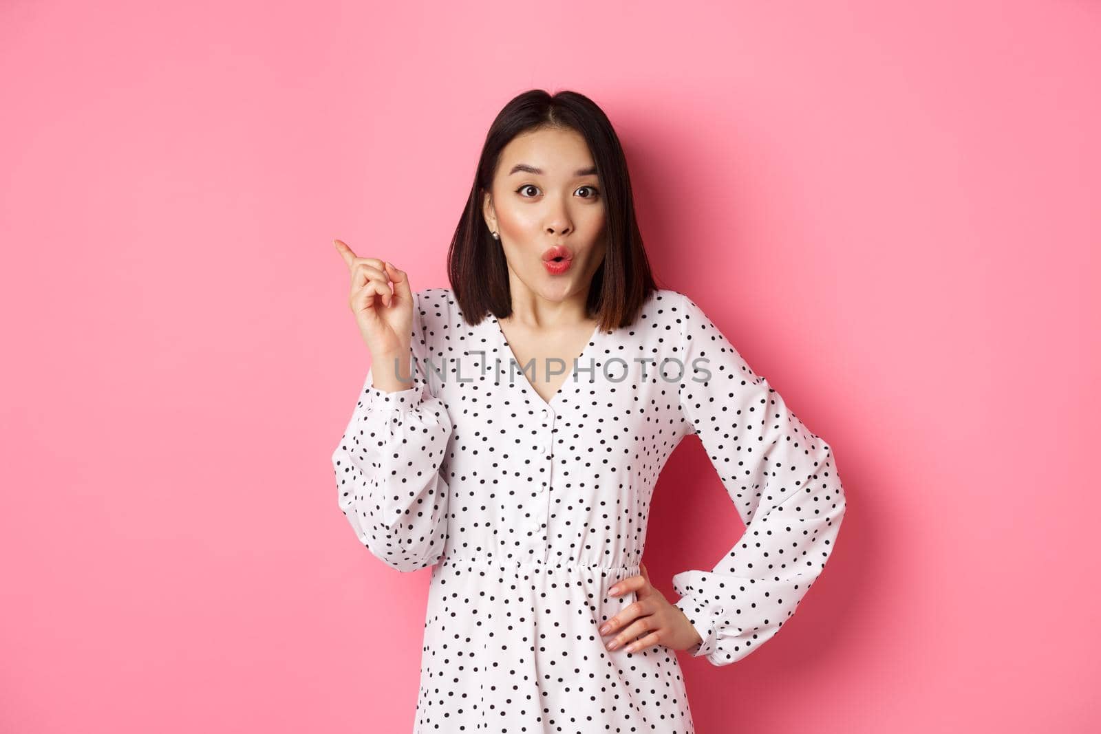 Excited asian female model showing promo offer, pointing at upper left corner and staring at camera amazed, wearing trendy spring dress, standing over pink background.