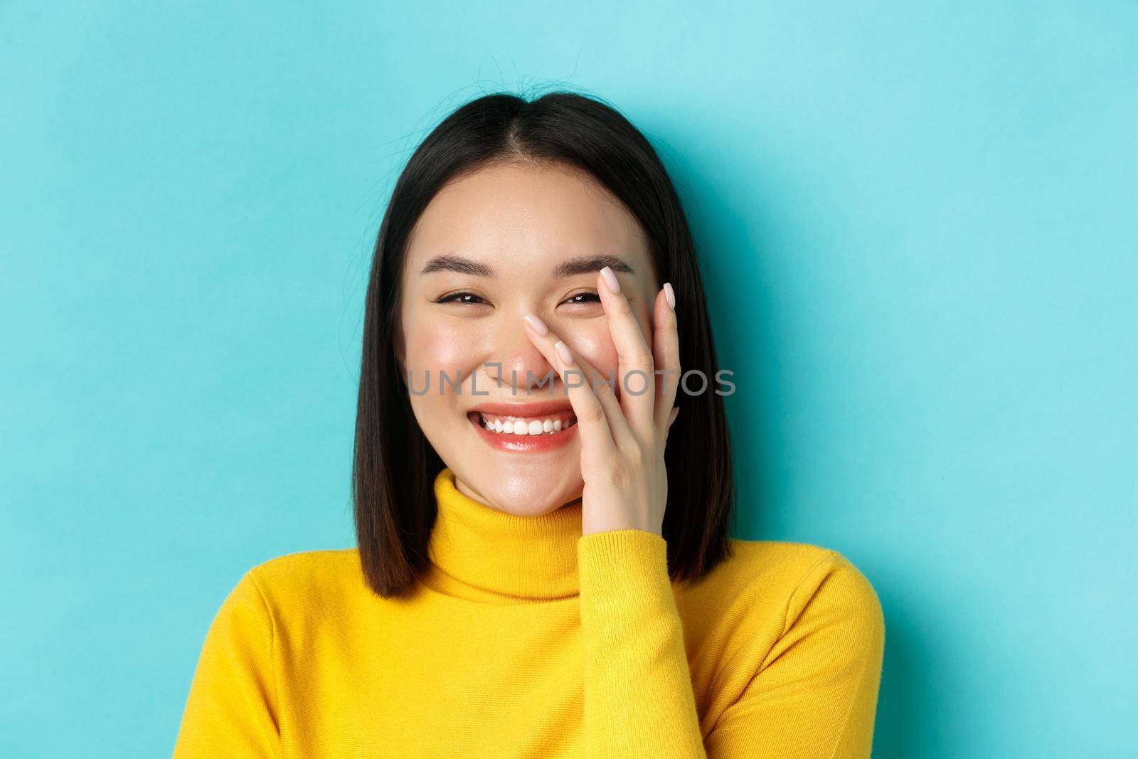Beauty and makeup concept. Close up of beautiful asian woman blushing and laughing, touching glowing healthy skin, smiling happy, standing over blue background.