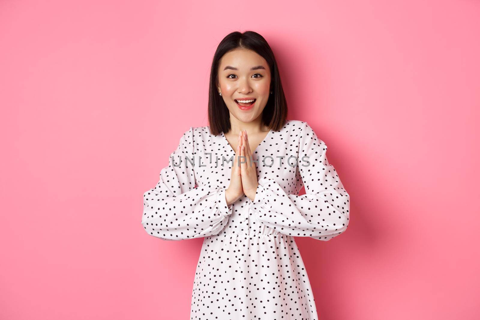 Beautiful asian woman thanking you, holding hands together in appreciation gesture, smiling happy at camera, standing grateful over pink background by Benzoix