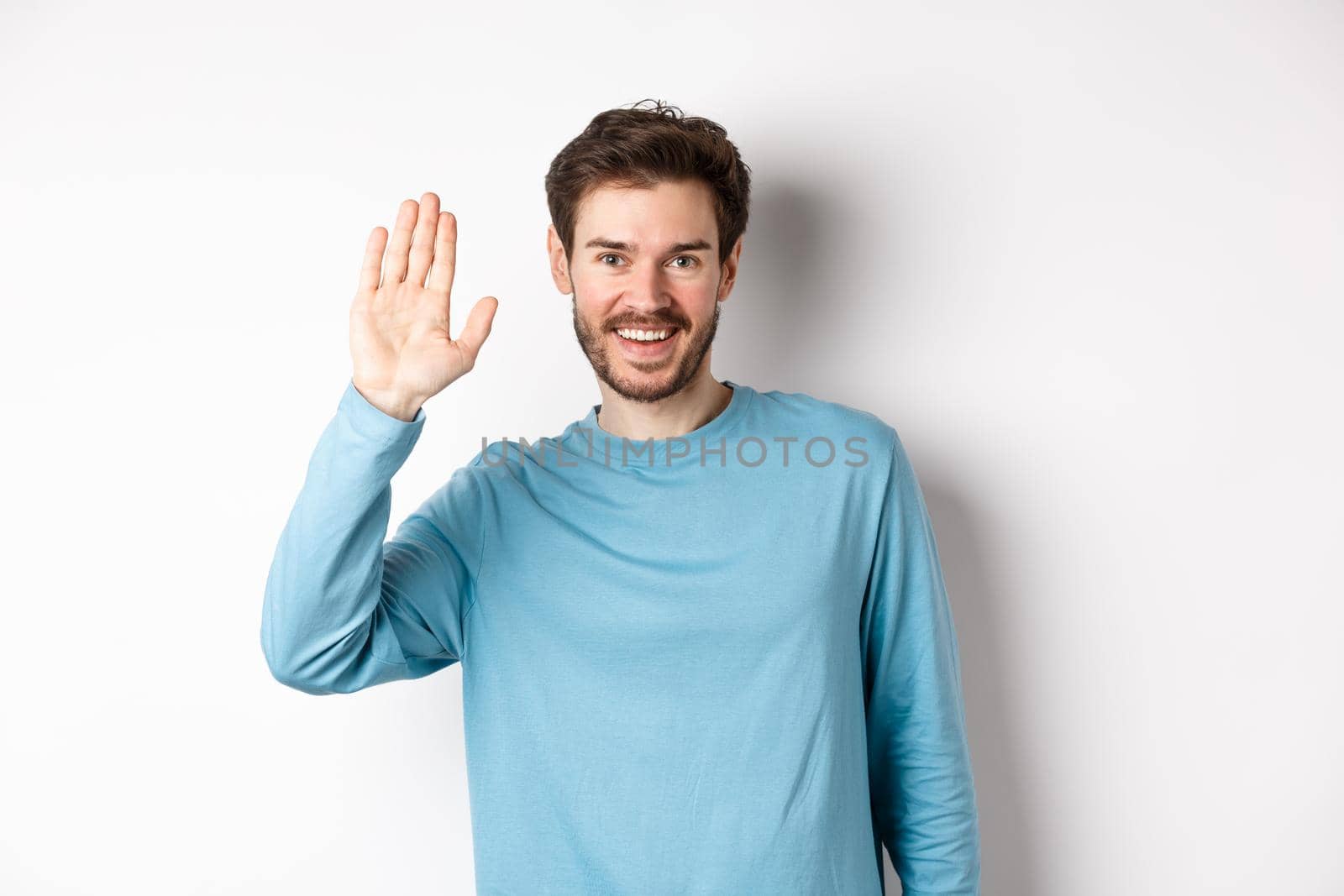 Cheerful young man with beard saying hello, looking friendly and waving hand to greet you, standing over white background by Benzoix