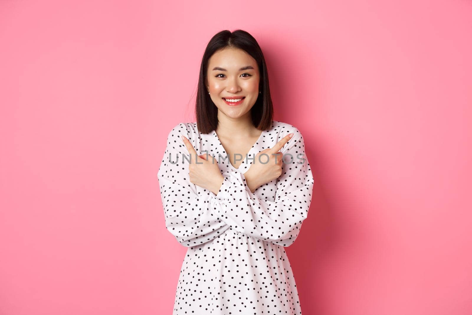 Beautiful asian girl in romantic dress pointing fingers sideways, showing two variants on shopping, smiling at camera, standing against pink background by Benzoix