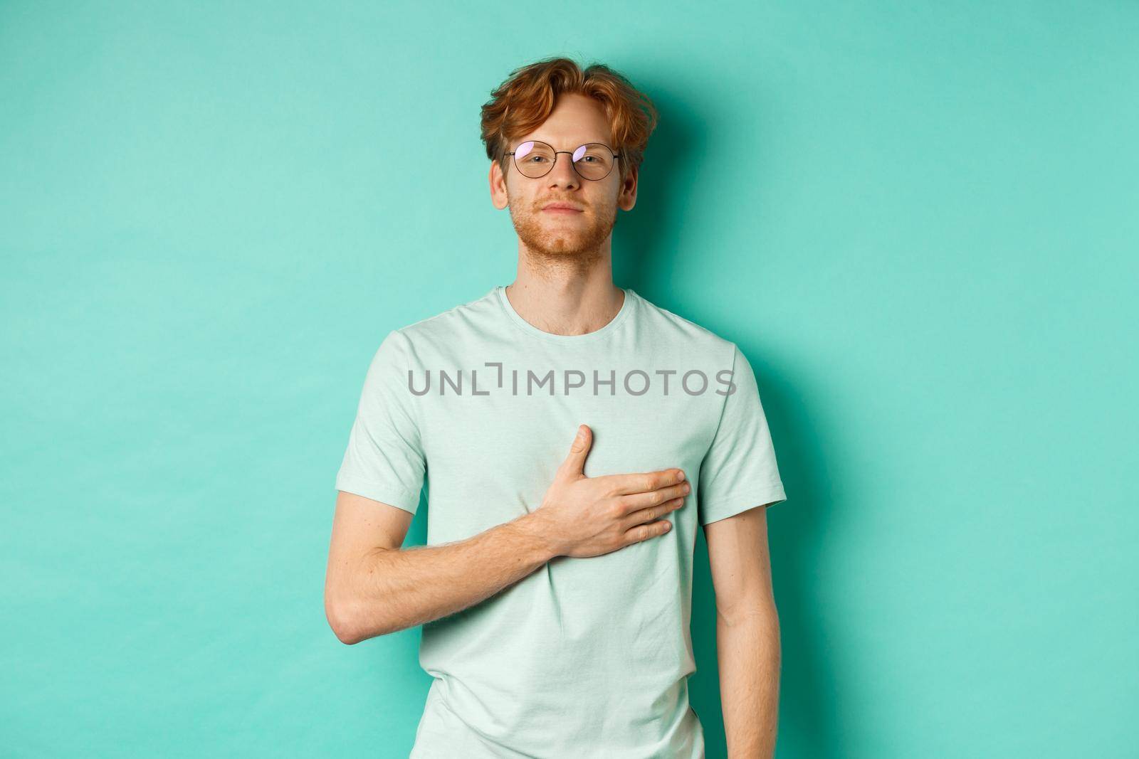 Proud young redhead man in glasses holding hand on heart, listening national anthem with respect, standing in t-shirt against turquoise background.