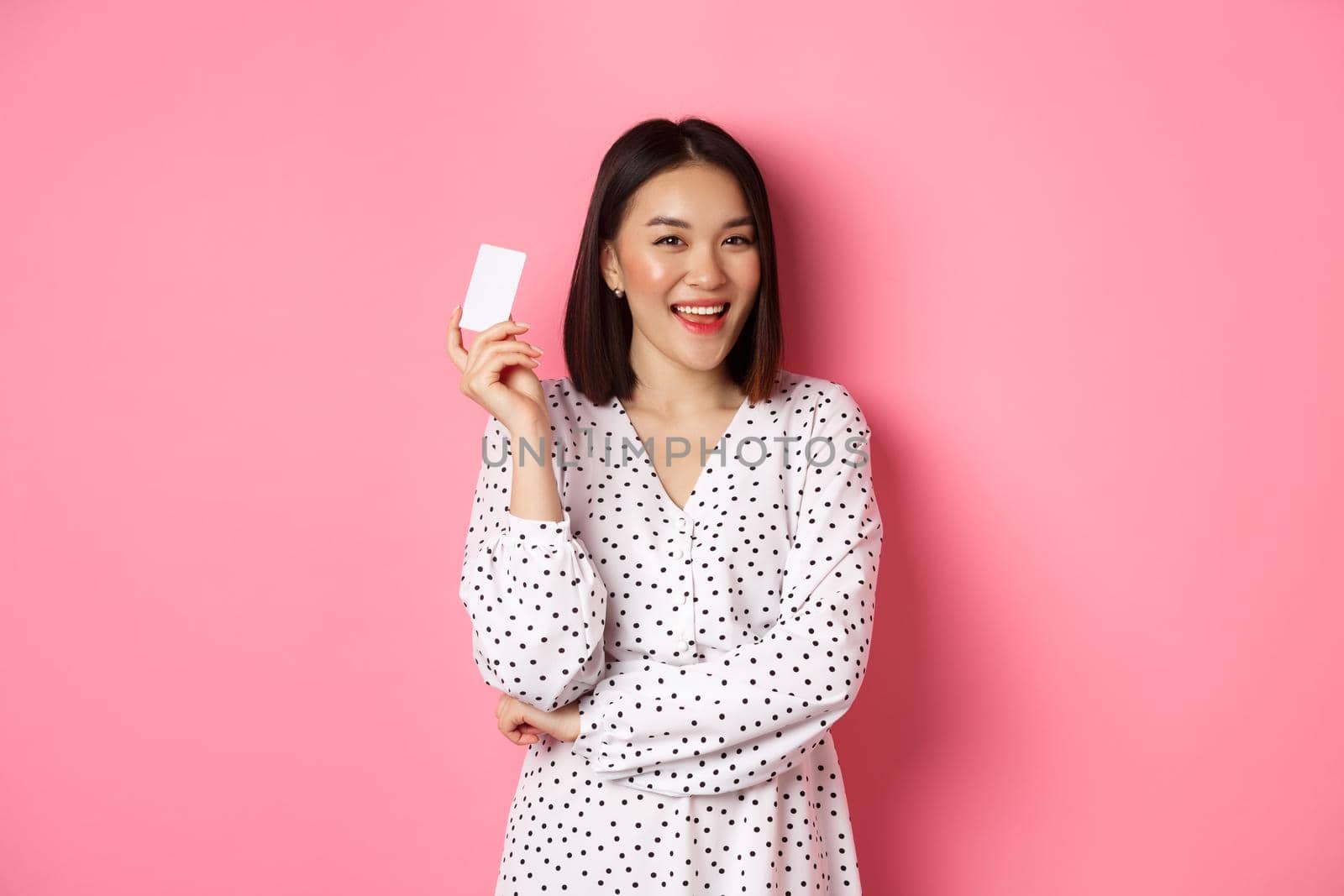 Shopping concept. Confident and happy asian woman holding credit card and smiling satisfied, standing against pink background by Benzoix