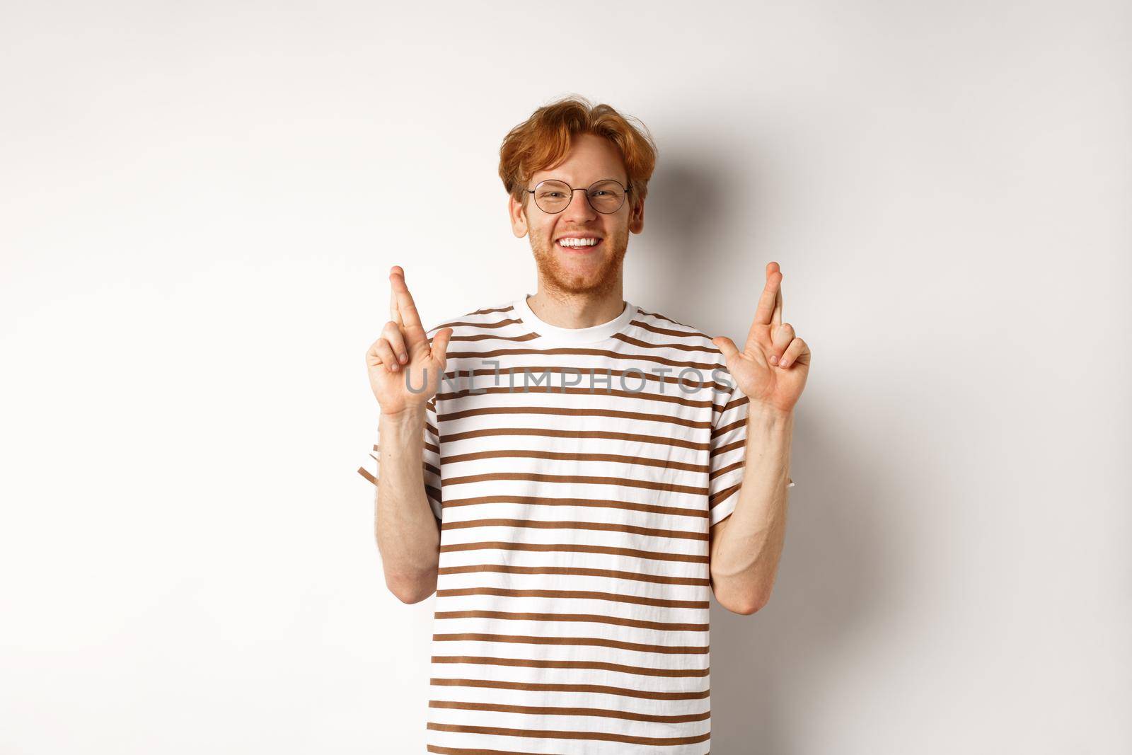 Hopeful and optimistic man with ginger hair and glasses having hope, making wish and cross fingers for good luck, smiling at camera, white background.