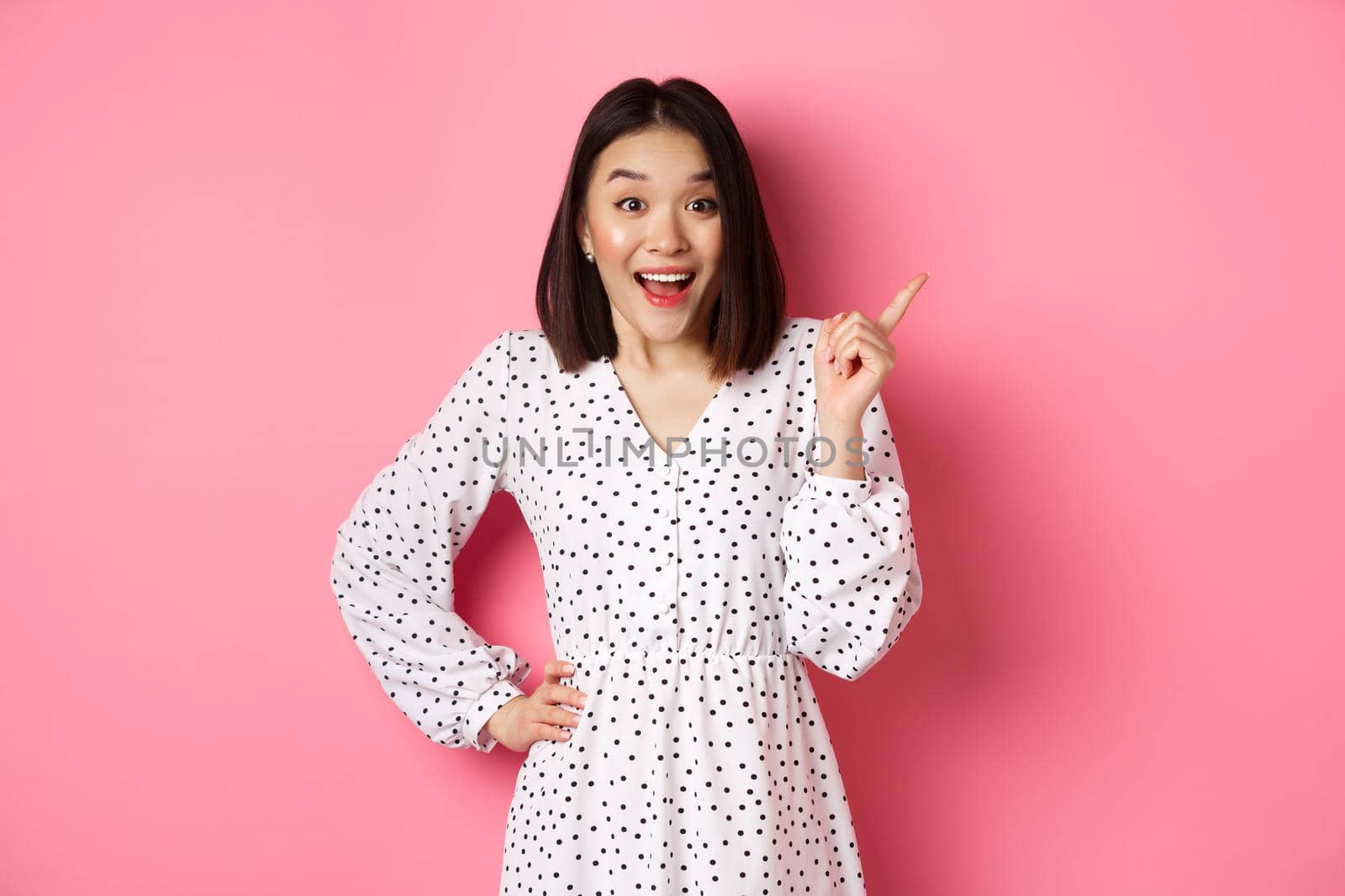 Excited pretty asian girl pointing at upper right corner, gasping amazed and looking at camera, discuss store promo offer, standing over pink background by Benzoix