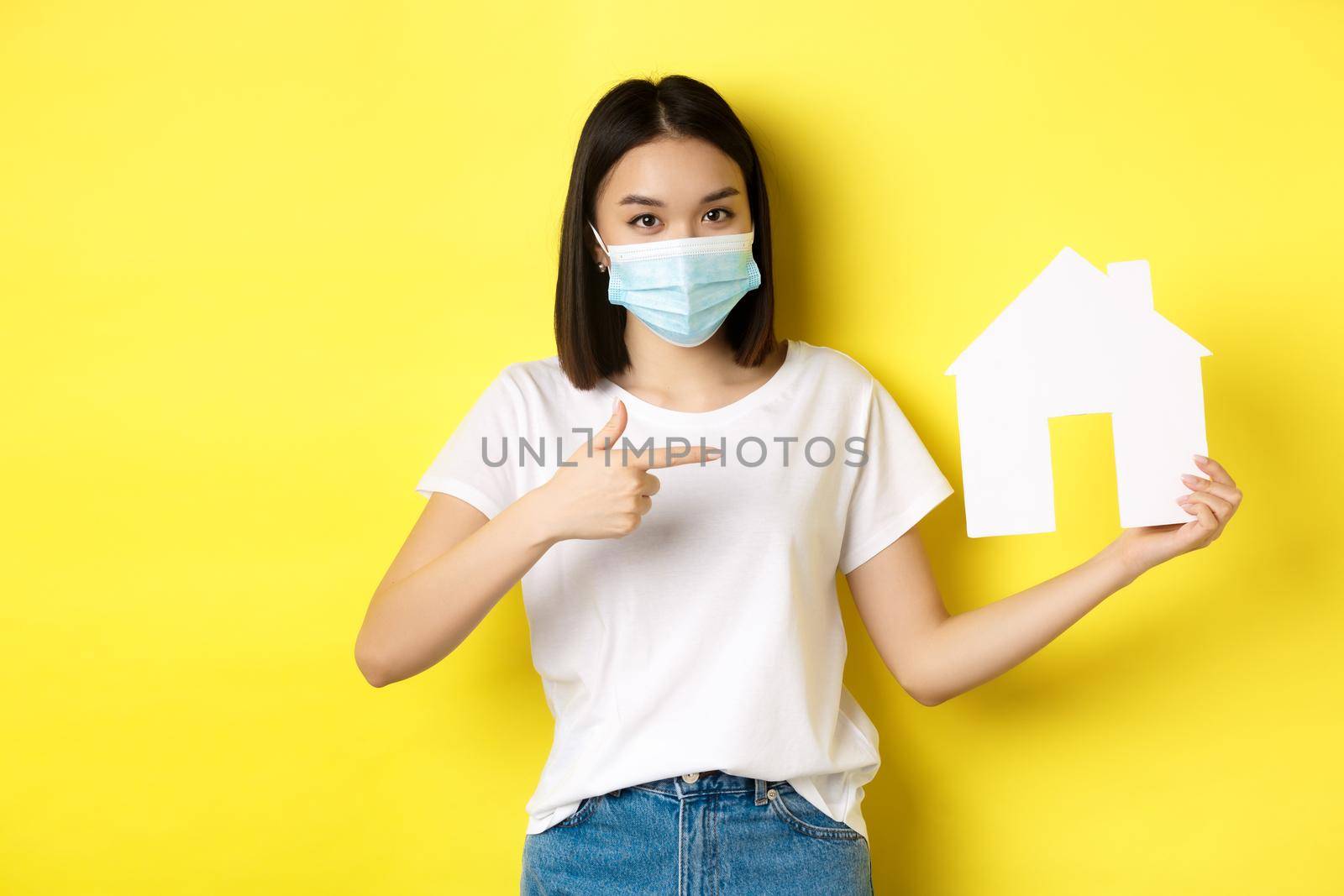 Covid-19, pandemic and real estate concept. Cheerful asian woman smiling in medical mask, showing paper house cutout, recommend agency for buying property.