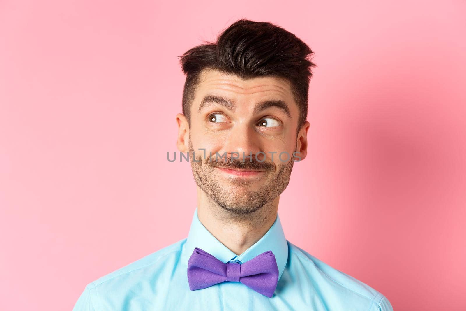 Close-up of handsome smiling guy with moustache, looking left at logo with pleased face, standing on pink background. Copy space