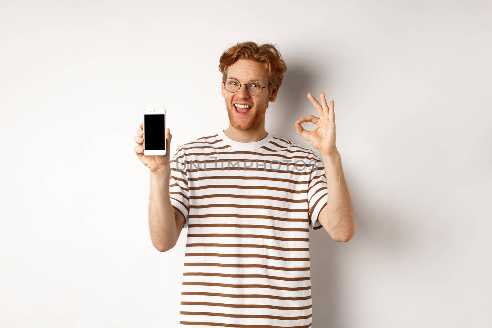 Technology and e-commerce concept. Young man with red hair showing okay sign and blank smartphone screen, praising awesome app, standing over white background by Benzoix