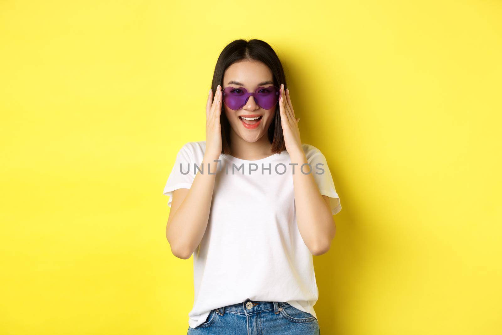 Fashion and lifestyle concept. Stylish asian woman trying new sunglasses in heart-shape, going on vacation, smiling happy at camera, standing over yellow background.