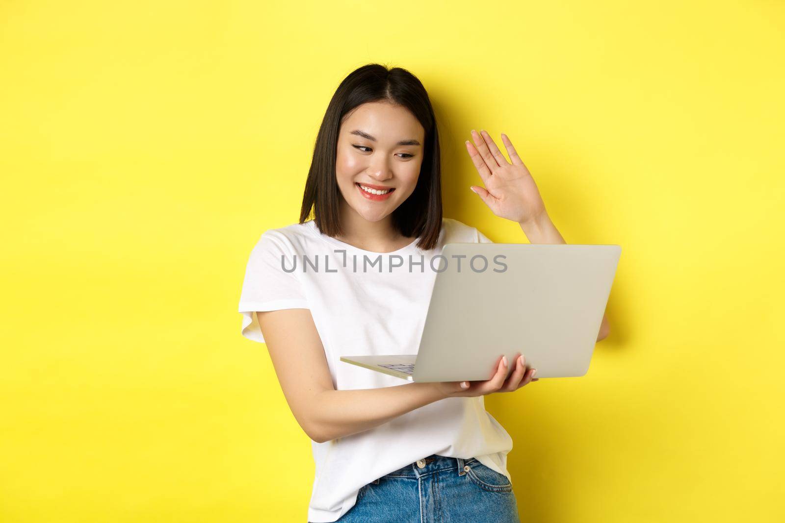 Happy asian woman video chat on laptop, waiving hand at computer camera and saying hello, standing over yellow background by Benzoix