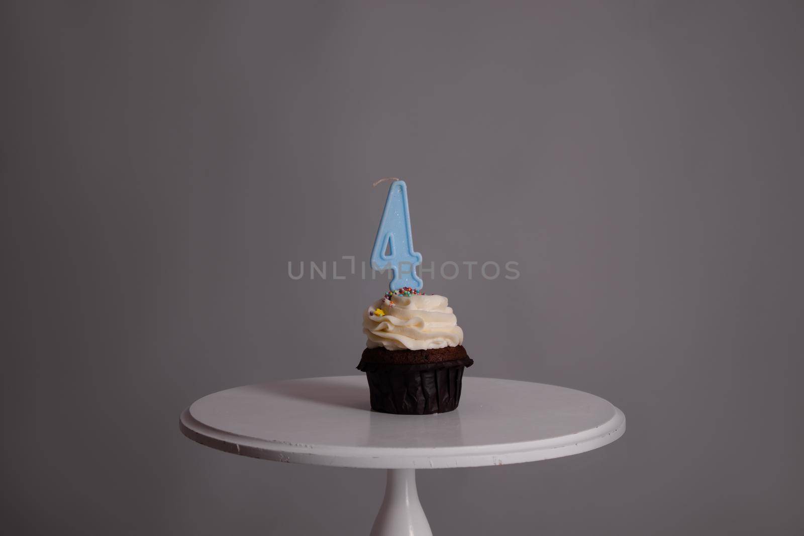 tasty chocolate cupcake with white topping cream and candle in shape of number four 4. on a white stand on a gray background. sweets, bakery. party food. dessert.