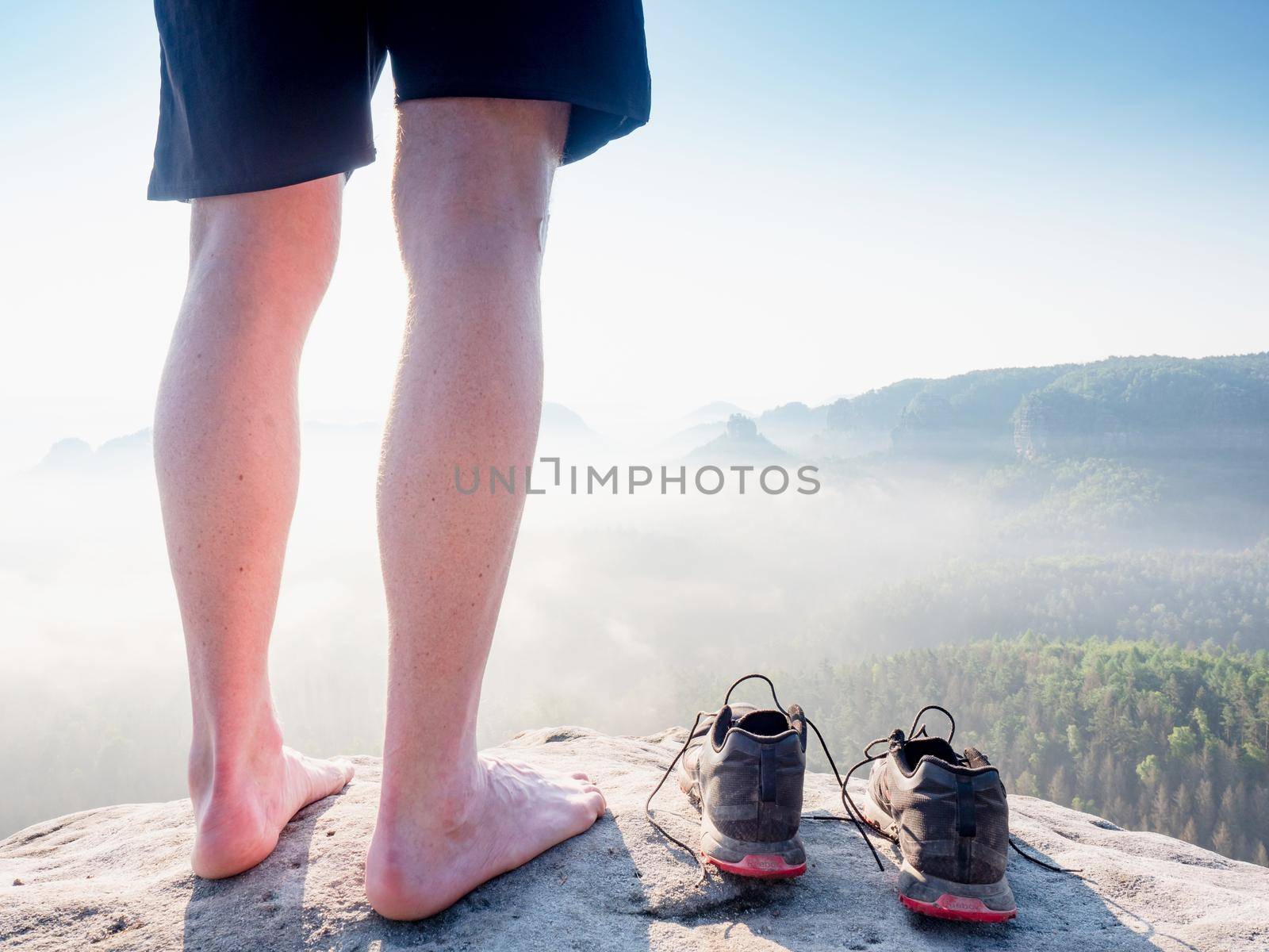 Slim barefoot legs with hairy calves of runner stay at to removed sweaty running shoes on a rocky edge above a long deep vally in nature park.