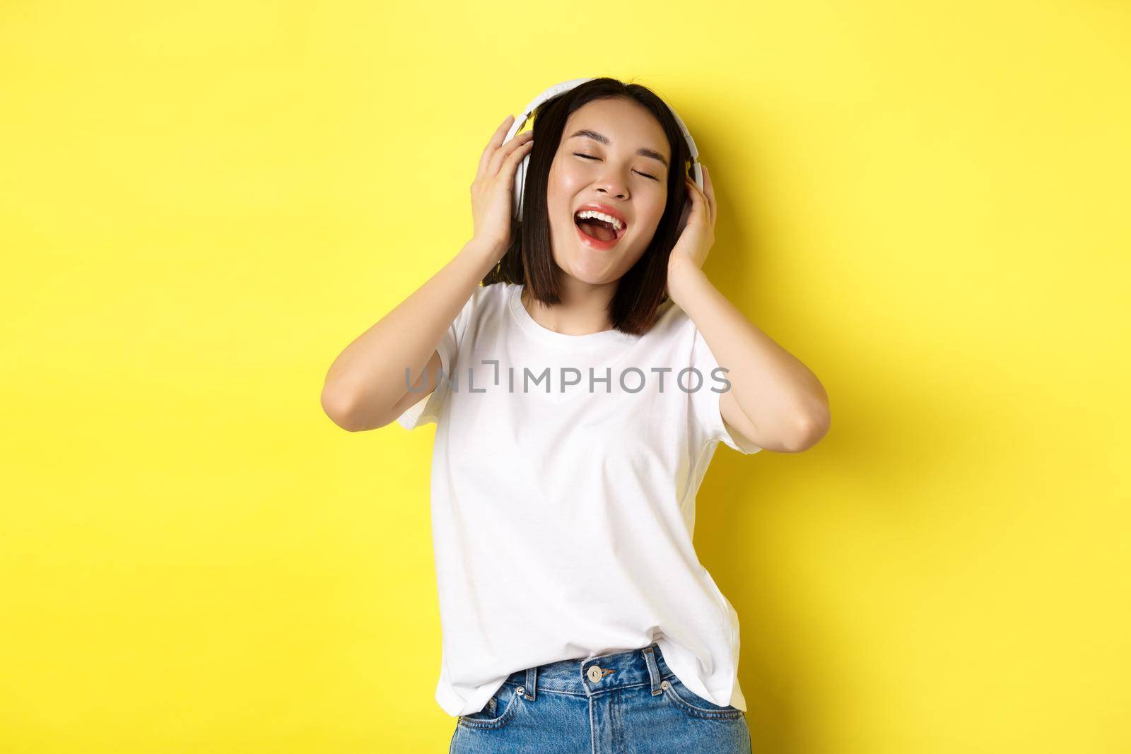 Attractive asian woman enjoying listening music in wireless headphones, smiling pleased and singing along, yellow background by Benzoix