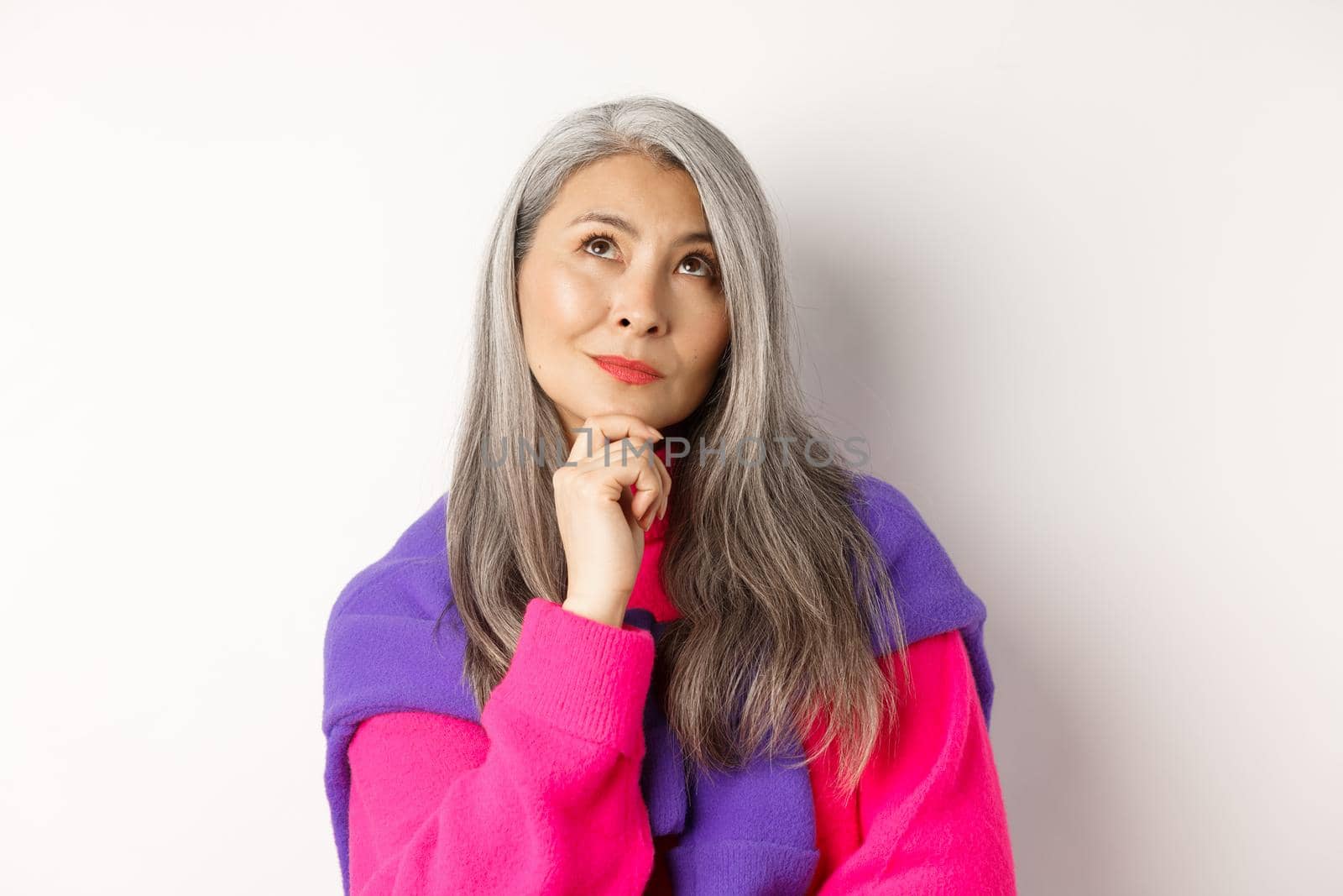 Close up of thoughtful asian female with grey hair, looking up and thinking, making decision, standing over white background by Benzoix