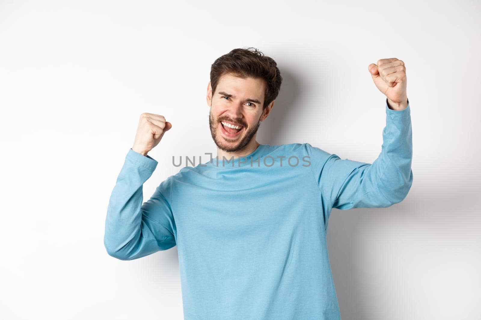 Image of happy young man triumphing, feeling like champion, winning and screaming in cheer, standing over white background.