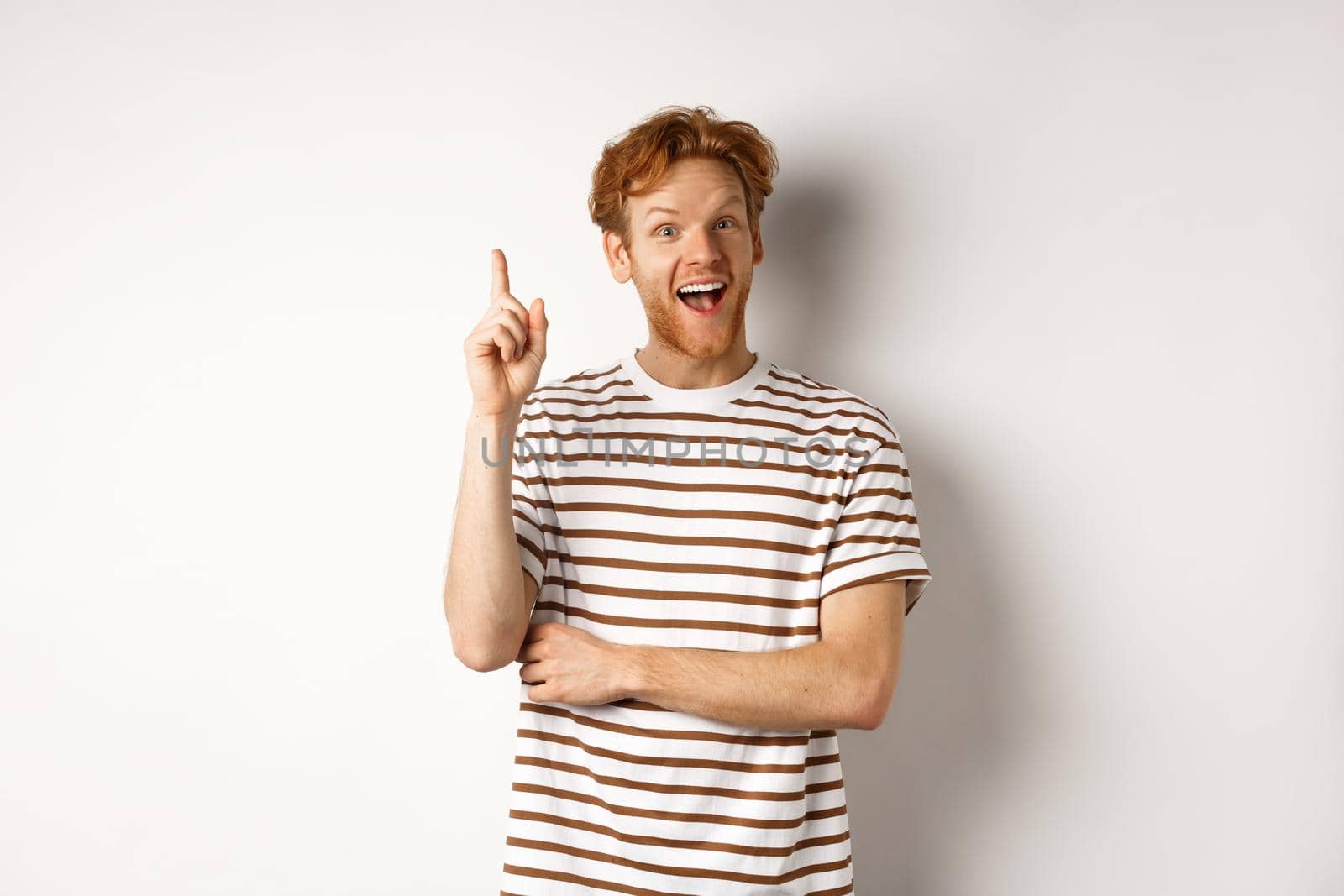 Young man with red hair having an idea, raising finger to say suggestion, standing happy over white background.