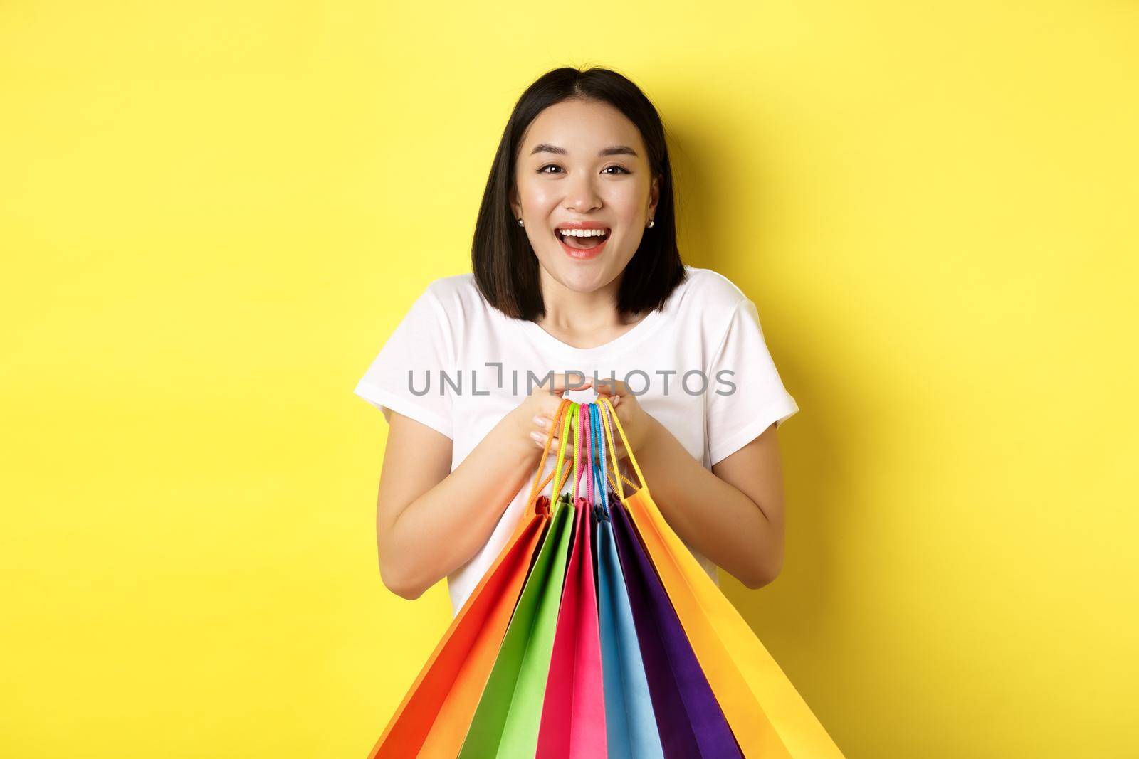 Happy asian female shopper smiling and holding colorful shopping bags, standing against yellow background.