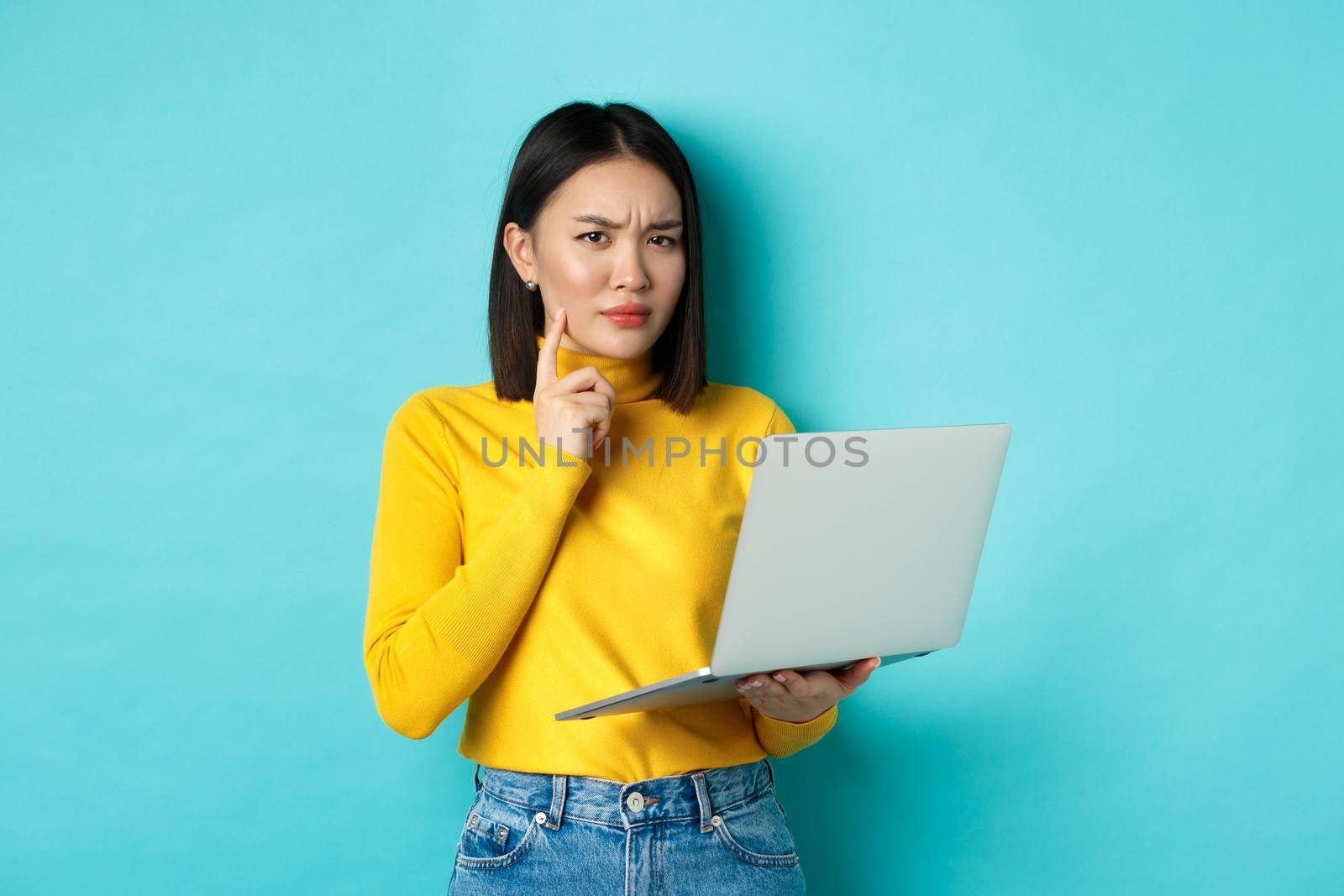 Serious looking asian woman working on laptop and thinking, frowning at camera, solving problem at work, standing over blue background by Benzoix