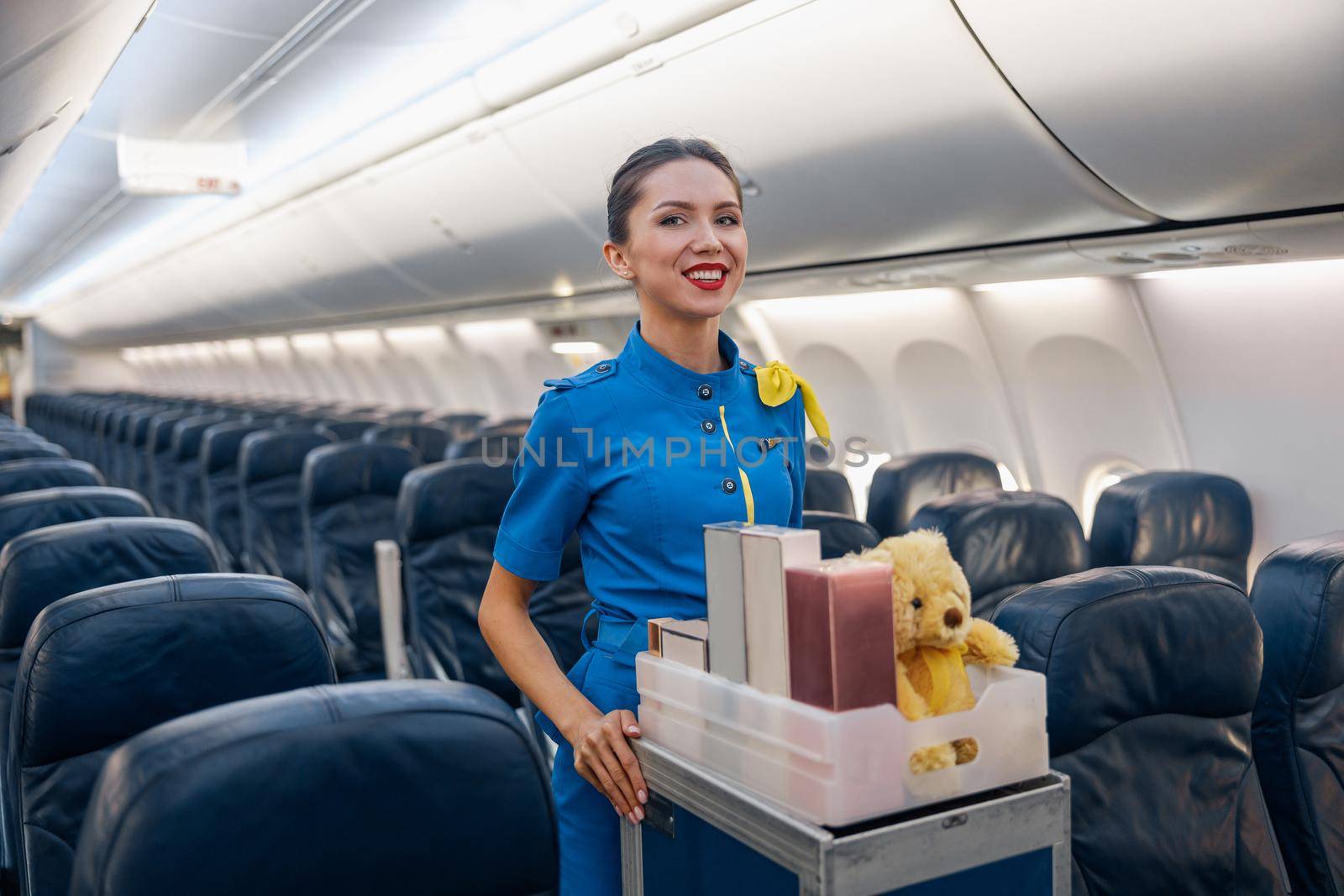Cheerful female air hostess in bright blue uniform smiling at camera while leading trolley cart with gifts through empty plane aisle. Travel, service, transportation, airplane concept