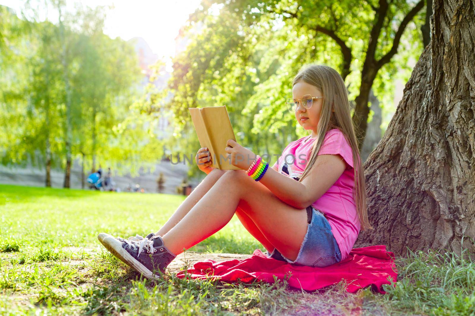 Girl child in glasses reading book in the park, on the grass near the tree