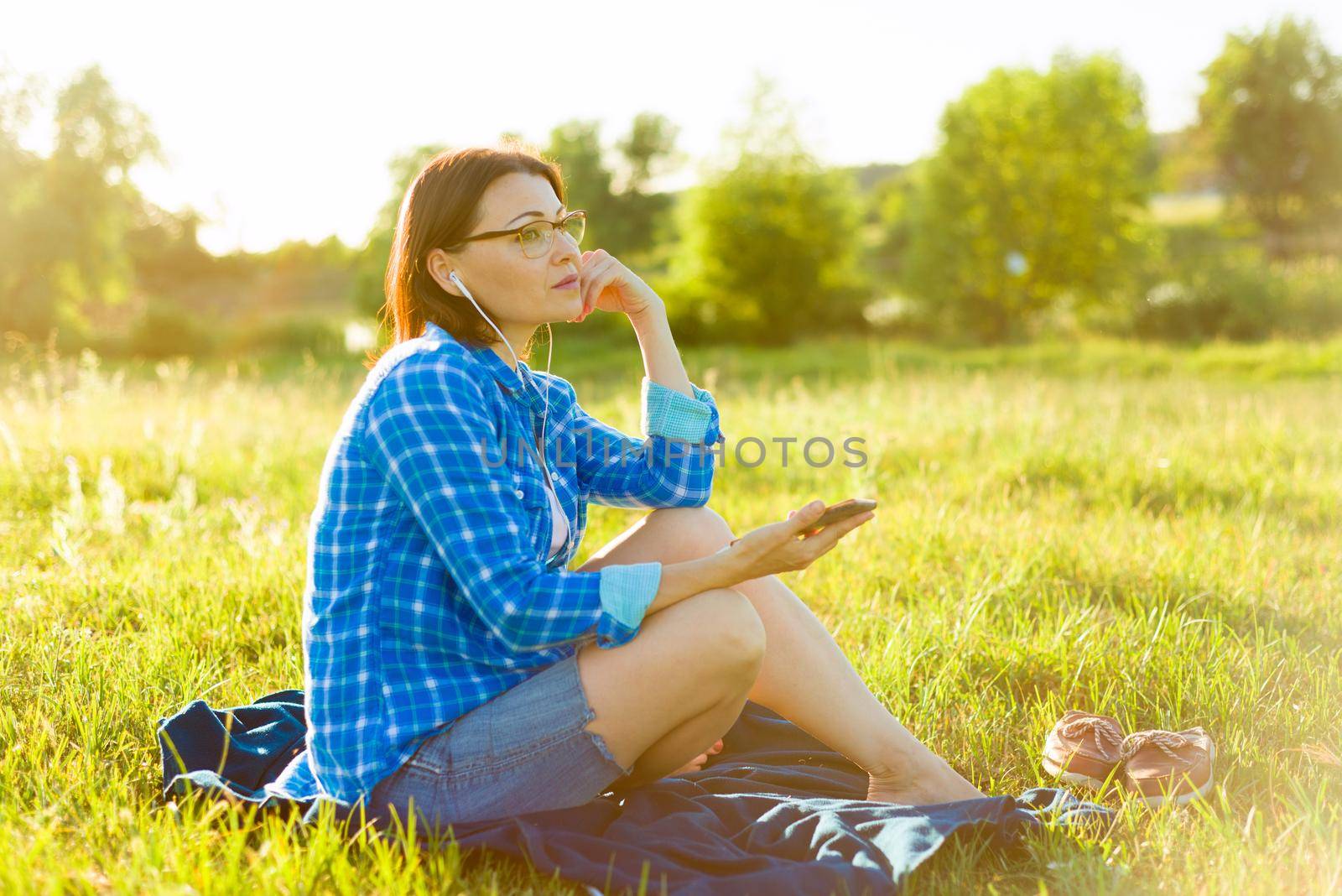 Mature woman listens to music, an audiobook on headphones, relaxes in nature. Background sunset, rustic landscapes, green meadow