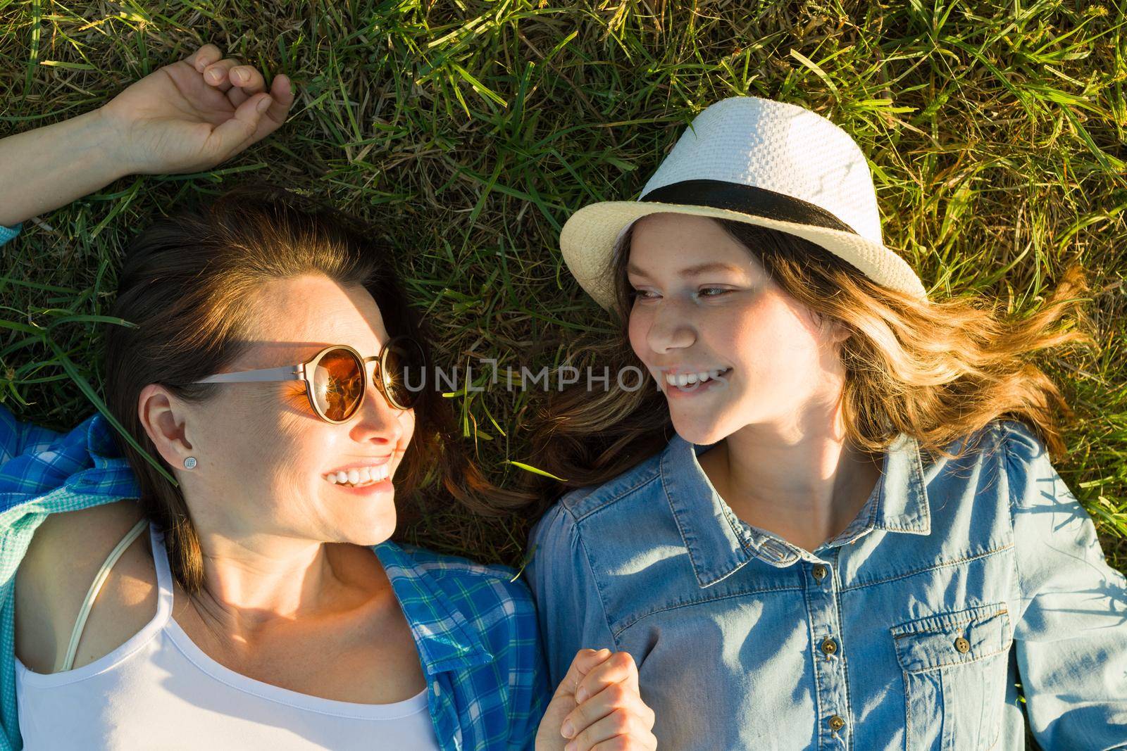 The parent and teenager, mom and 14 year old daughter are smiling lying on the green grass. view from above,