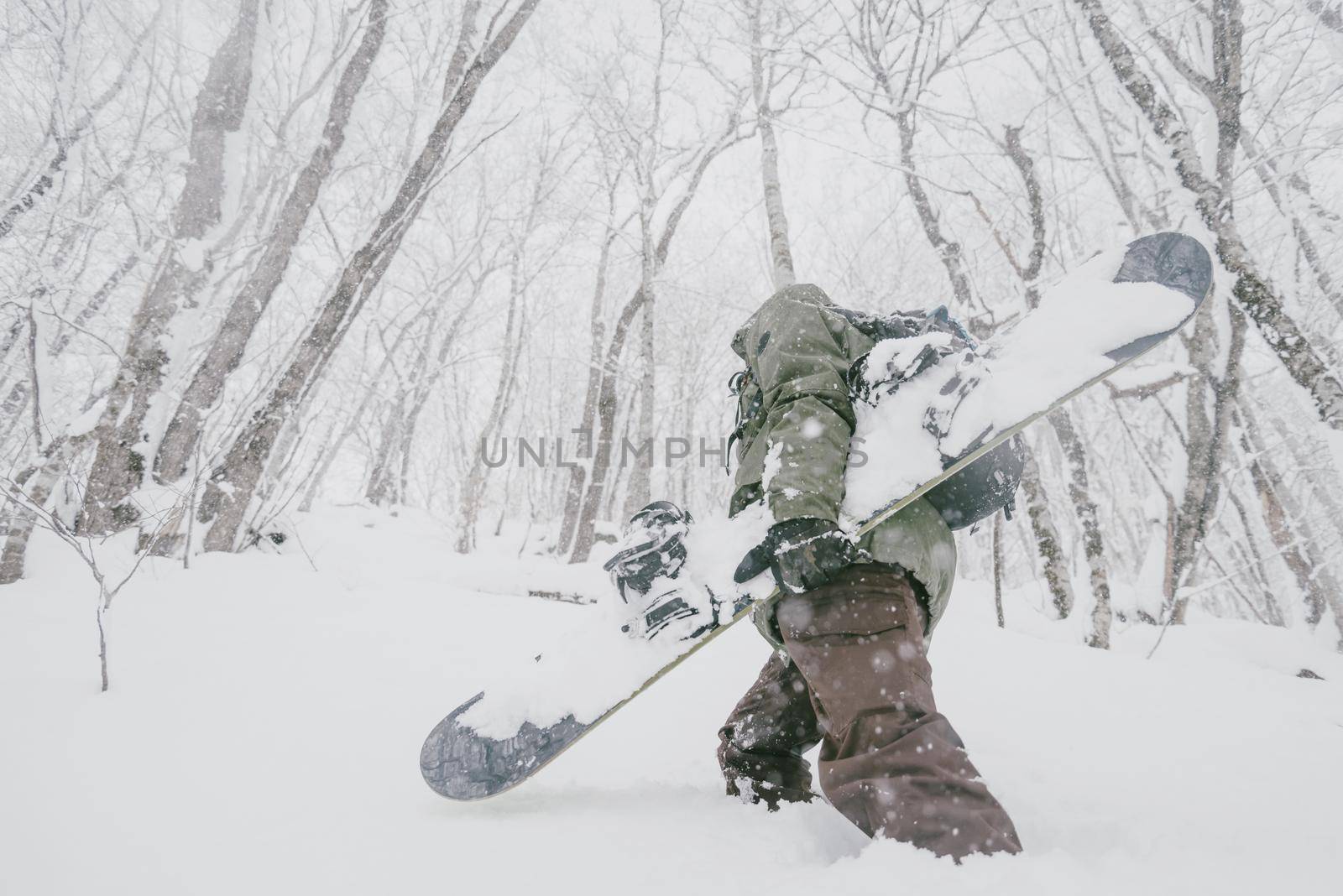 Unrecognizable freerider young man with snowboard walking in winter forest among snowdrifts