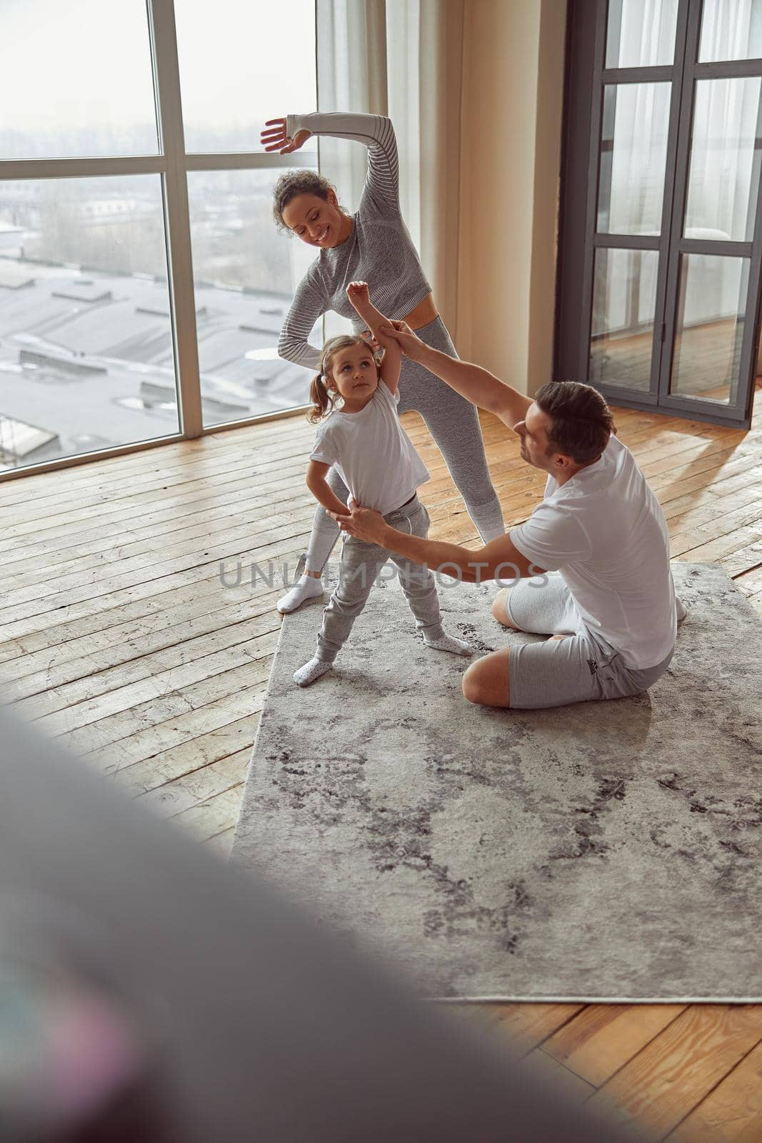 Top view of happy father and mother teaching their little daughter exercising in light living room