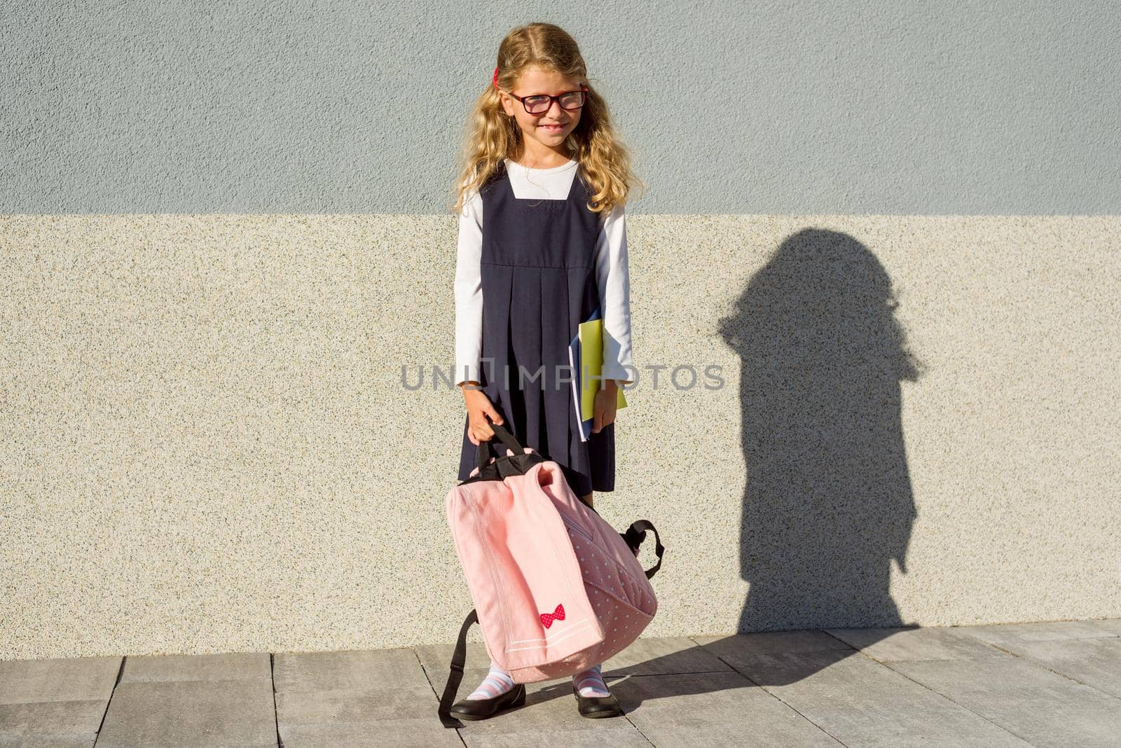 An elementary school student with notebooks in his hand. A little schoolgirl with a backpack near the building in the open air.