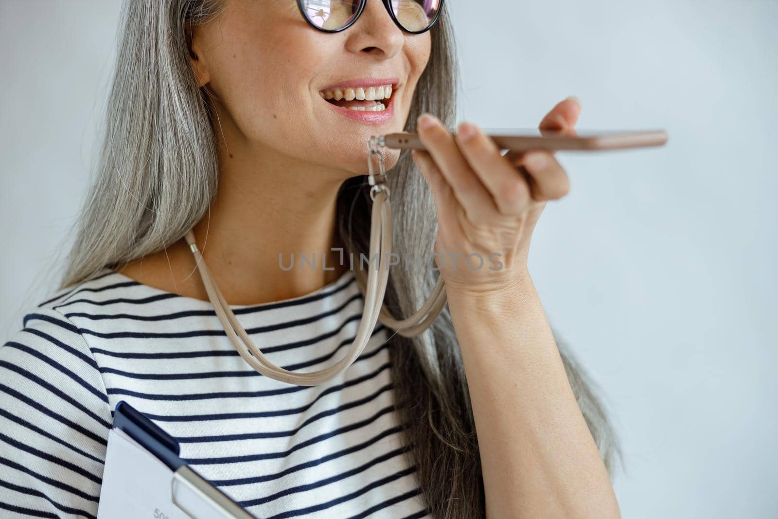 Positive mature lady with clipboard uses loudspeaker mode on mobile phone standing on light grey background in studio closeup