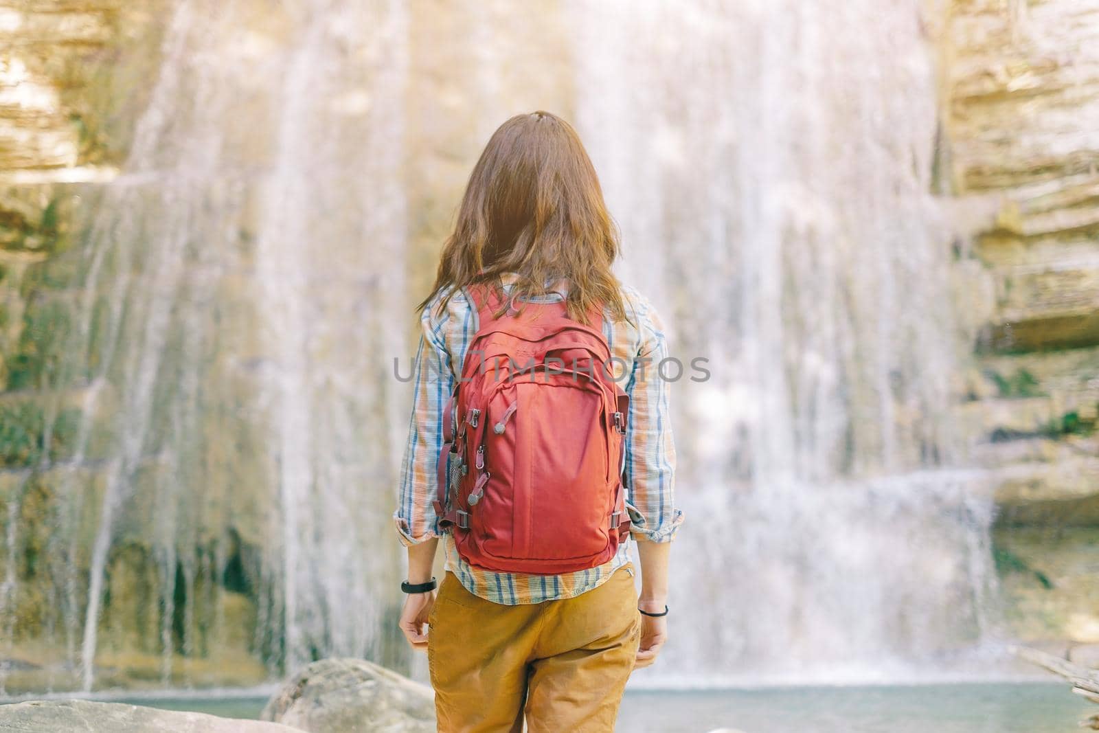 Unrecognizable hiker woman with backpack standing in front of waterfall, rear view.