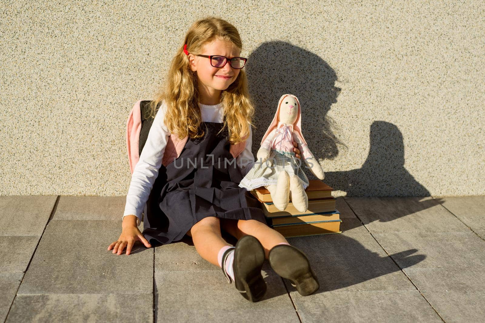 A cute schoolgirl of primary school is smiling, holding a toy. Girl with a backpack near the building outdoors. Leaving childhood and Back to school!
