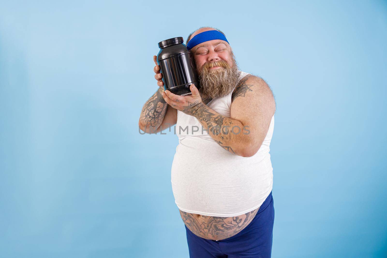 Positive middle aged man with overweight hugs large black bottle of sport nutrition standing on light blue background in studio