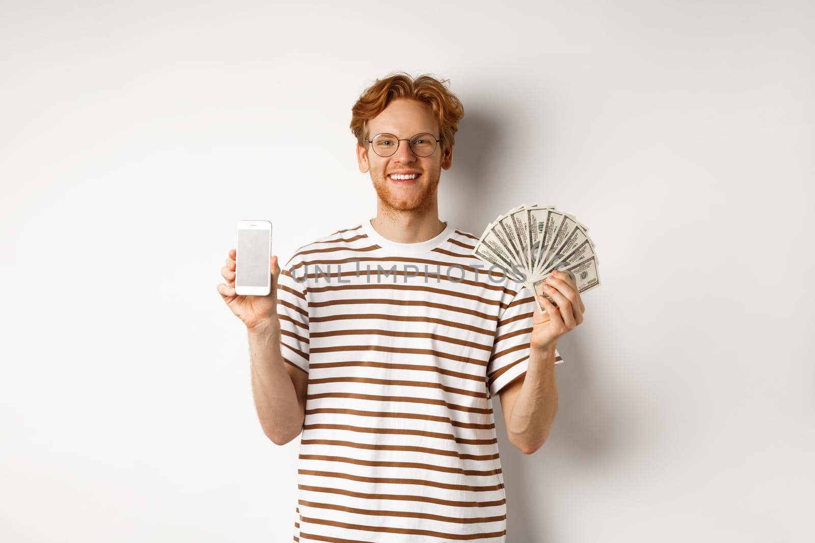 Smiling young redhead man in glasses showing smartphone blank screen and money, standing over white background by Benzoix