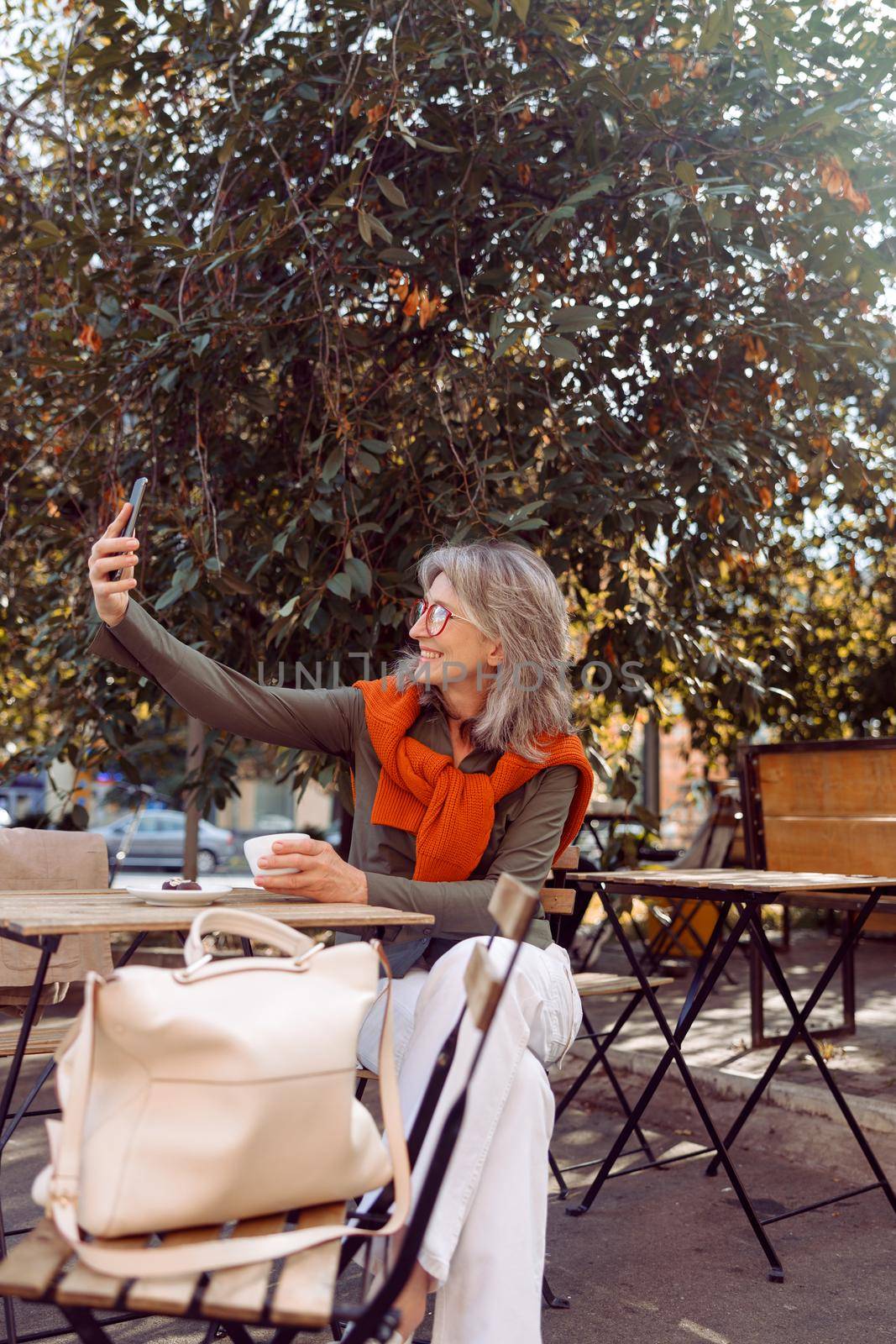 Pretty senior woman with glasses takes selfie with smartphone sitting at table on outdoors cafe terrace on autumn day