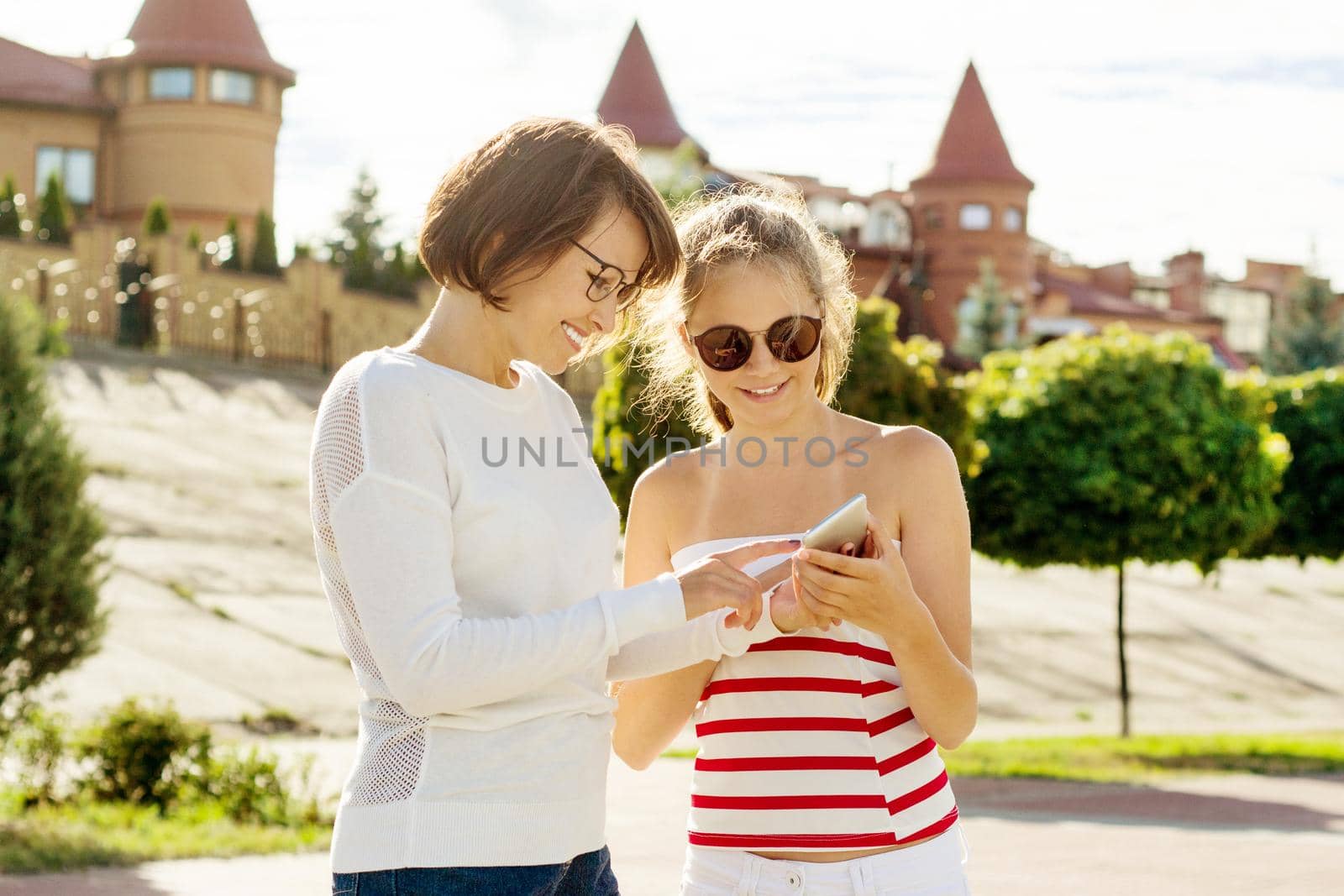 Mother and teen daughter portrait. Background summer city, mom and daughter are looking in smartphone.