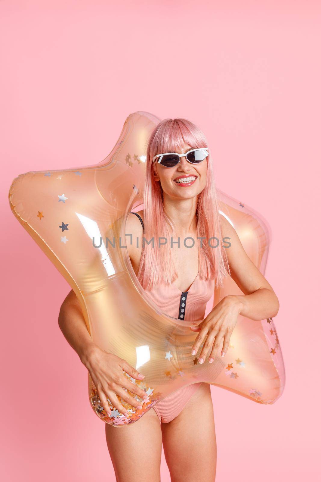 Joyful young woman with pink hair in swimsuit and sunglasses with inflatable star for swimming posing isolated over pink background by Yaroslav_astakhov