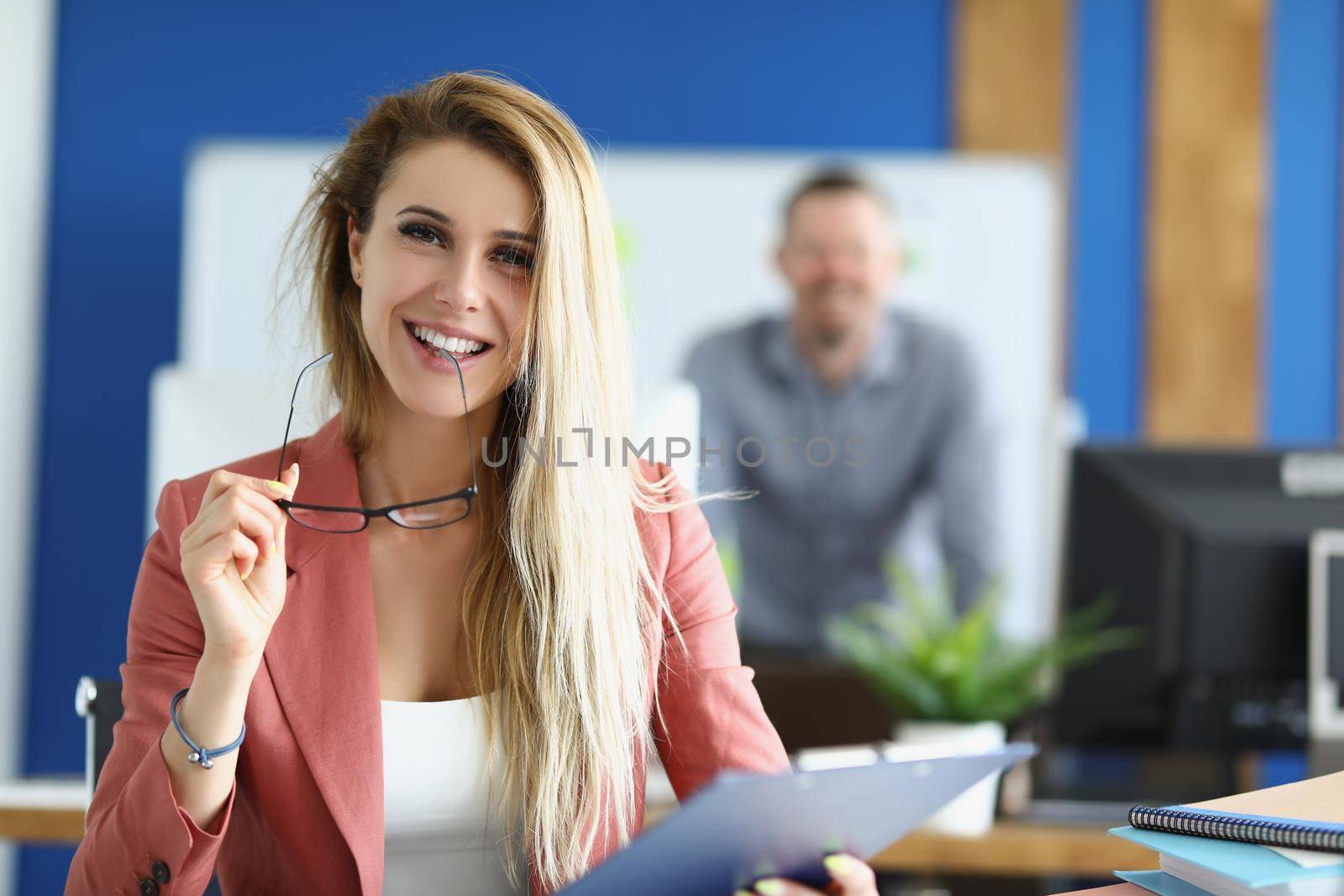 Portrait of cheerful optimistic female with clipboard, company dress code, office worker in suit. Perform daily tasks on workplace, woman employee, colleague on background. Business, career concept