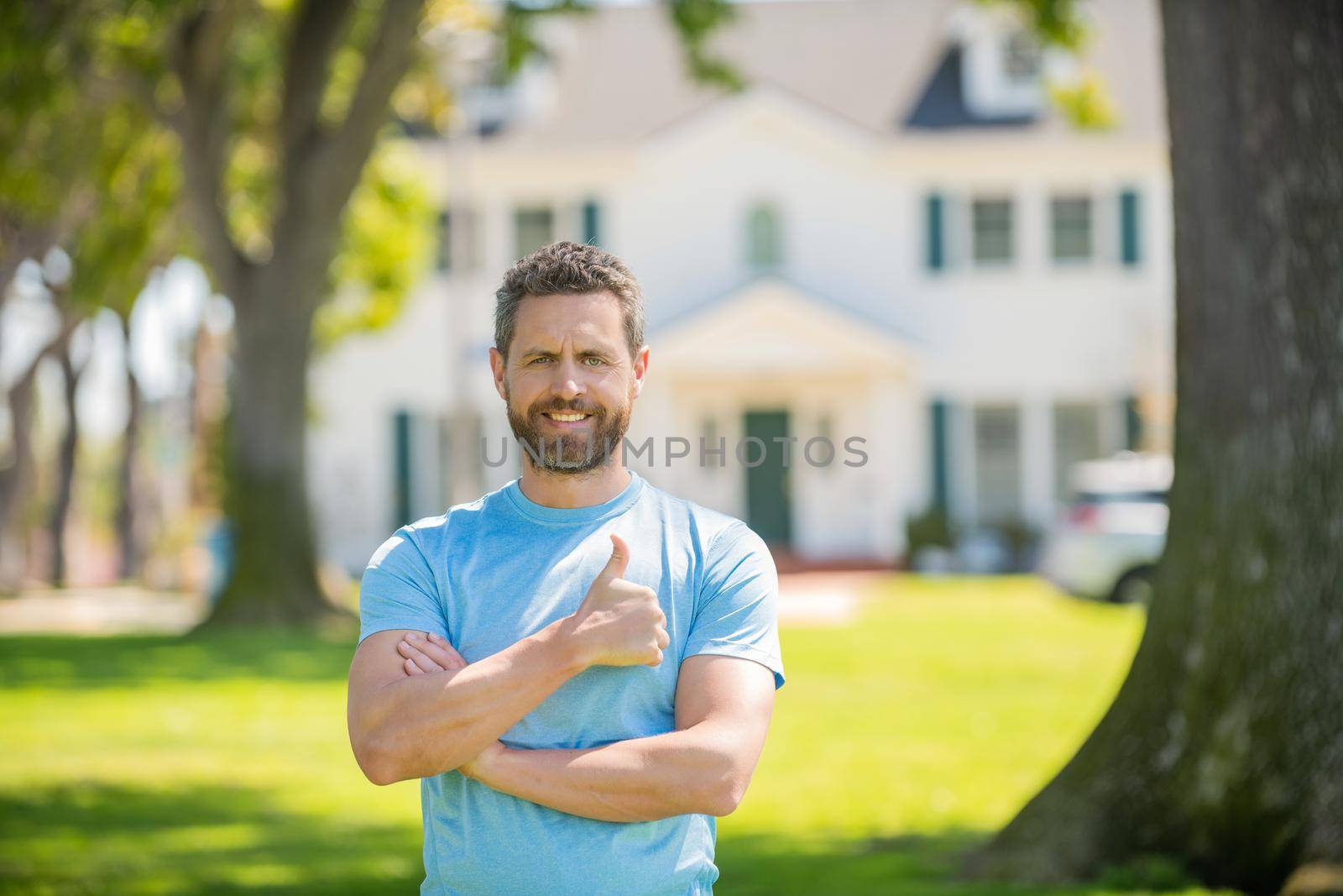 happy bearded man standing showing thumb upon house background, broker.