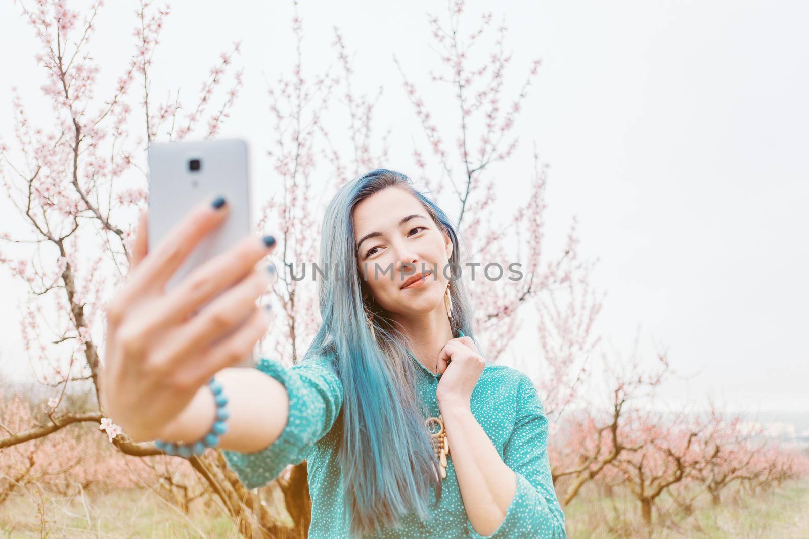 Beautiful young woman with blue hair taking selfie with smartphone in blossoming spring garden.