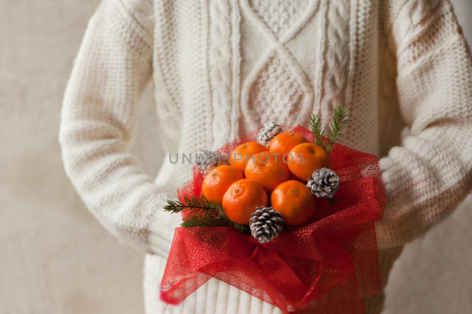 Woman hands in white sweater holding bouquet of mandarins and Christmas tree branches. New Year's edible bouquet of fruits. Gift for Christmas. DIY gift. useful gift made of fruits. decor of fruits.