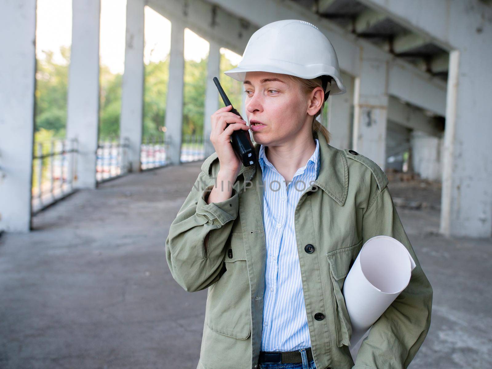 A female engineer in a white helmet looks at the construction of a building and speaks on a walkie-talkie. Architect holding a sketch of a building renovation plan