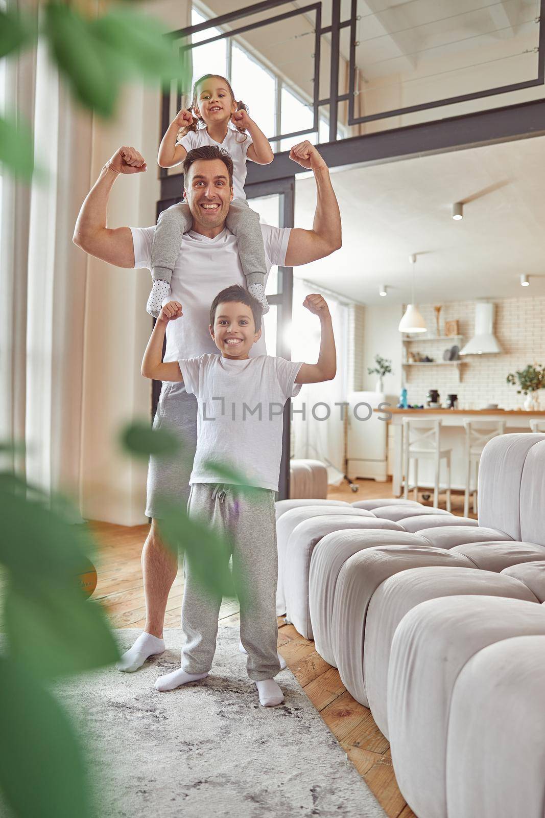 Full length portrait of cheerful man standing with son and daughter in living room and showing muscles