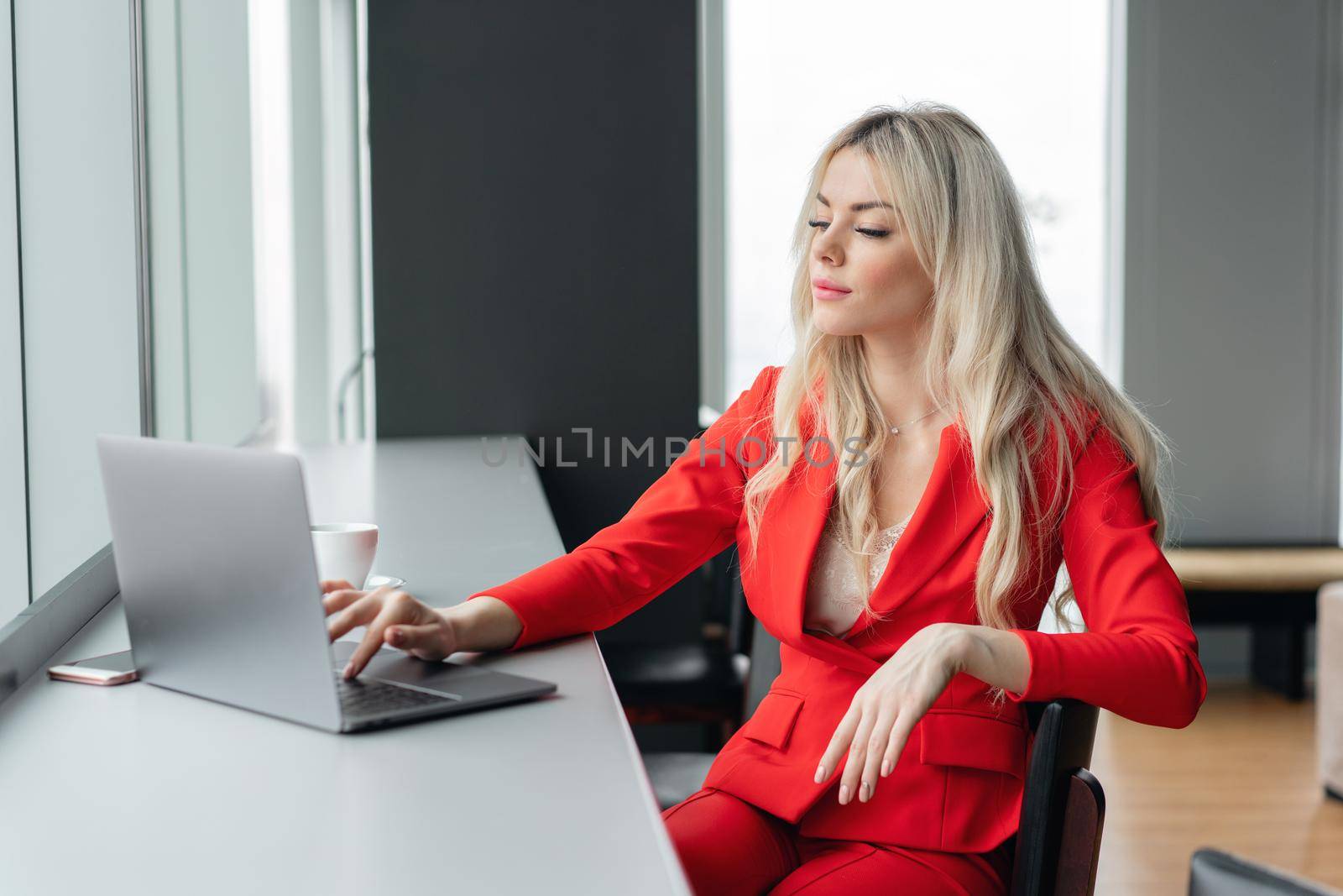 Woman in red coral business suit looks into the monitor screen. Young blonde businesswoman with modern gray laptop in the office in high floor in front of the window. Girl drinking coffee by Malkovkosta