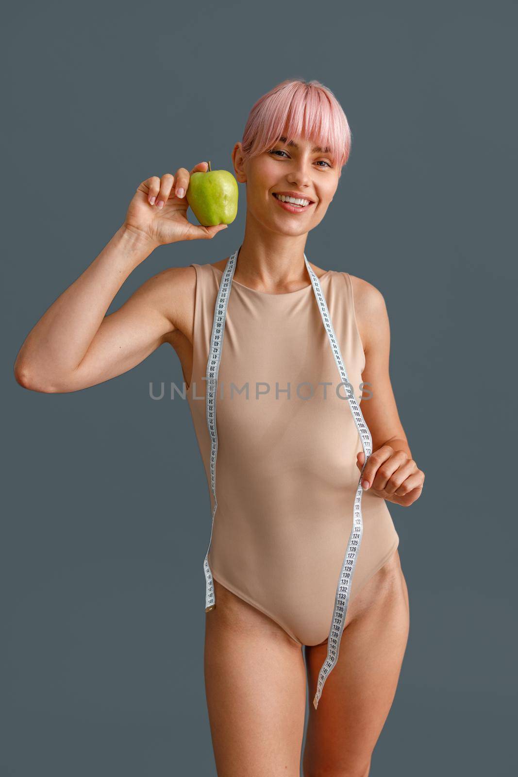 Smiling slim young woman in beige bodysuit holding green apple and measuring tape, standing isolated over gray studio background by Yaroslav_astakhov