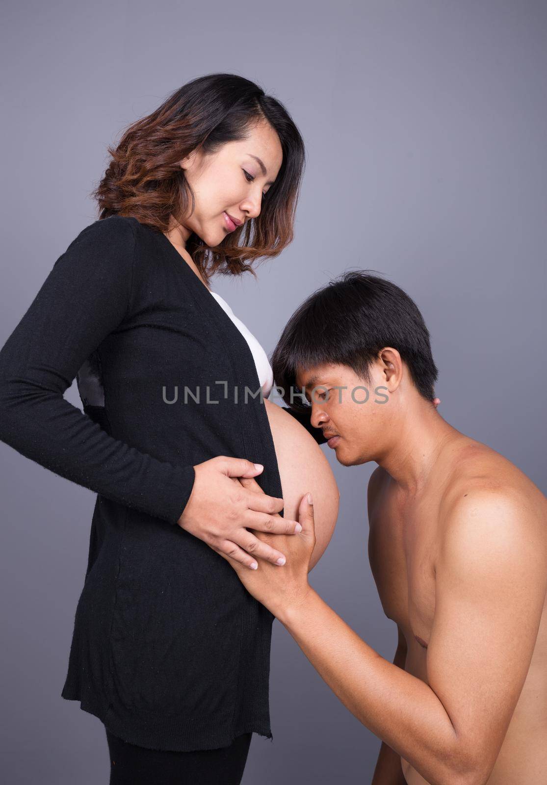 Young man kissing his pregnant wife's belly on gray background by geargodz