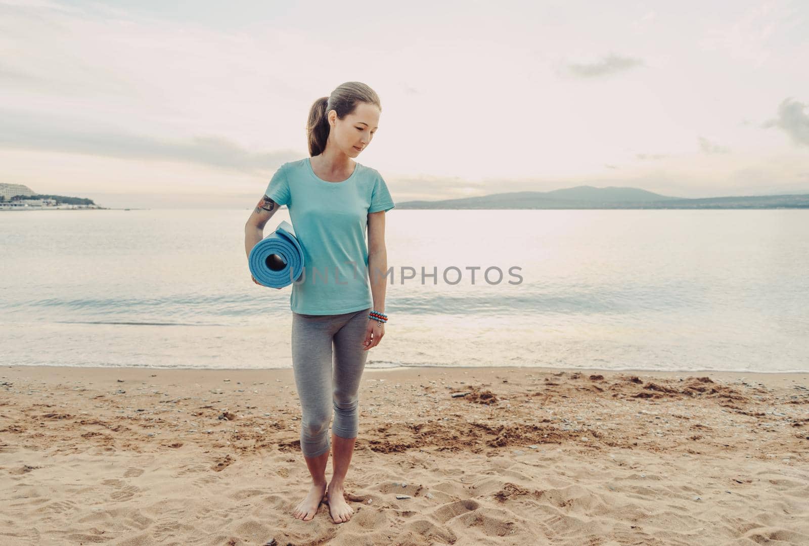 Young woman walking on sand beach with yoga mat on background of sea.