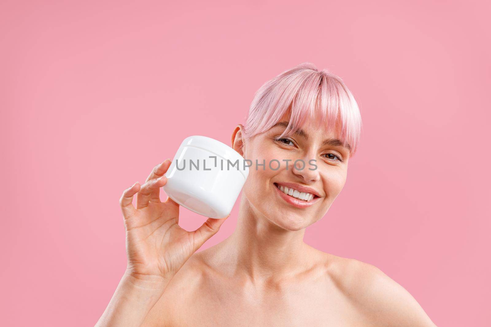 Portrait of smiling woman with pink hair holding white jar with moisturizing body lotion after shower isolated over pink background by Yaroslav_astakhov